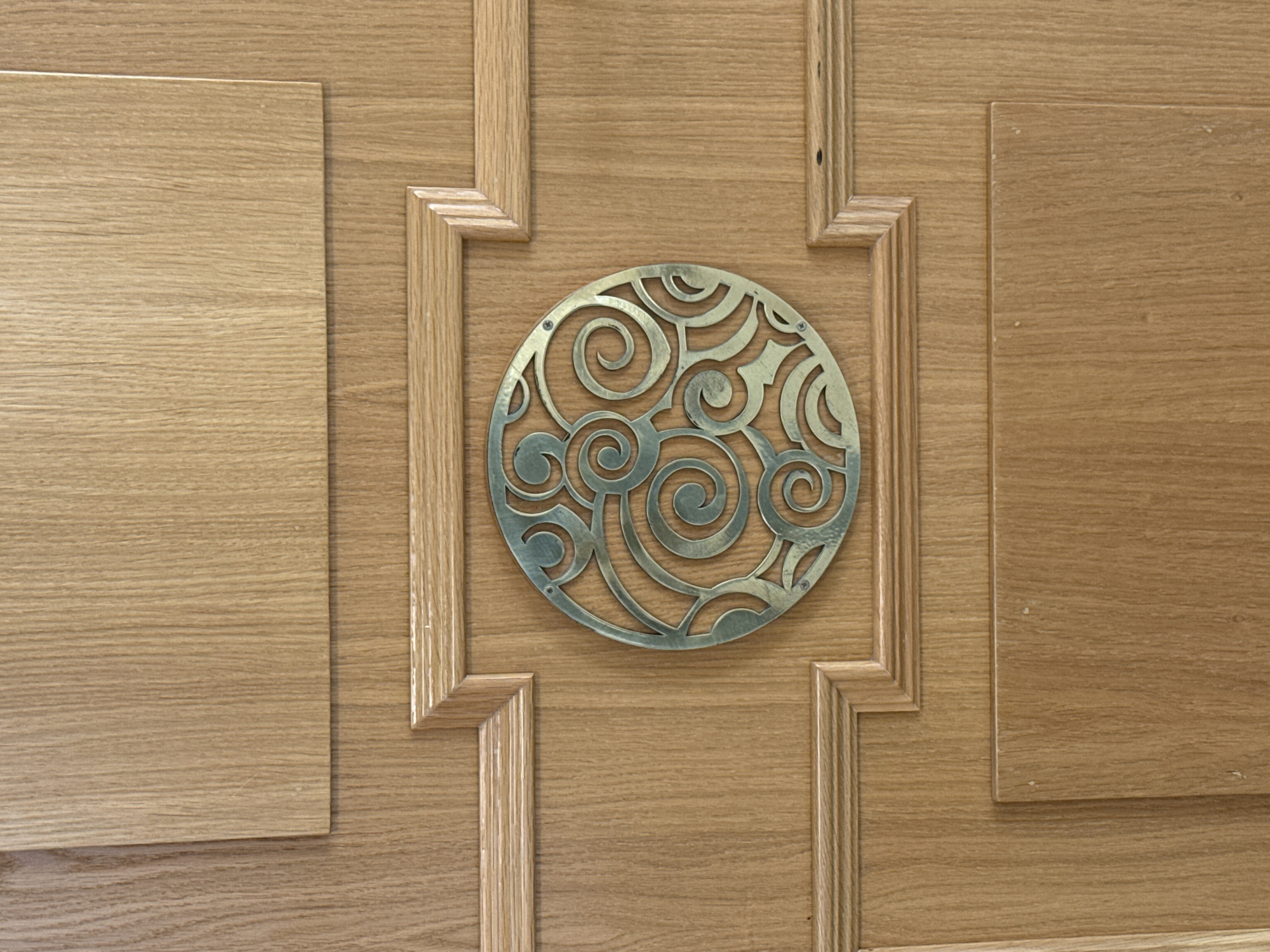 A close-up shot reveals a wooden door with a decorative, circular metal plate affixed to its center. The door appears to be made of light-colored wood with a subtle grain pattern. The design of the door includes rectangular panels. A circular metal plate is affixed with screws. The metal plate features an intricate, swirling design with several spirals. The image is well-lit, allowing clear visibility of the wood grain and metal details. There is no evidence to indicate the location of the image in the city of Votkinsk, Russia.