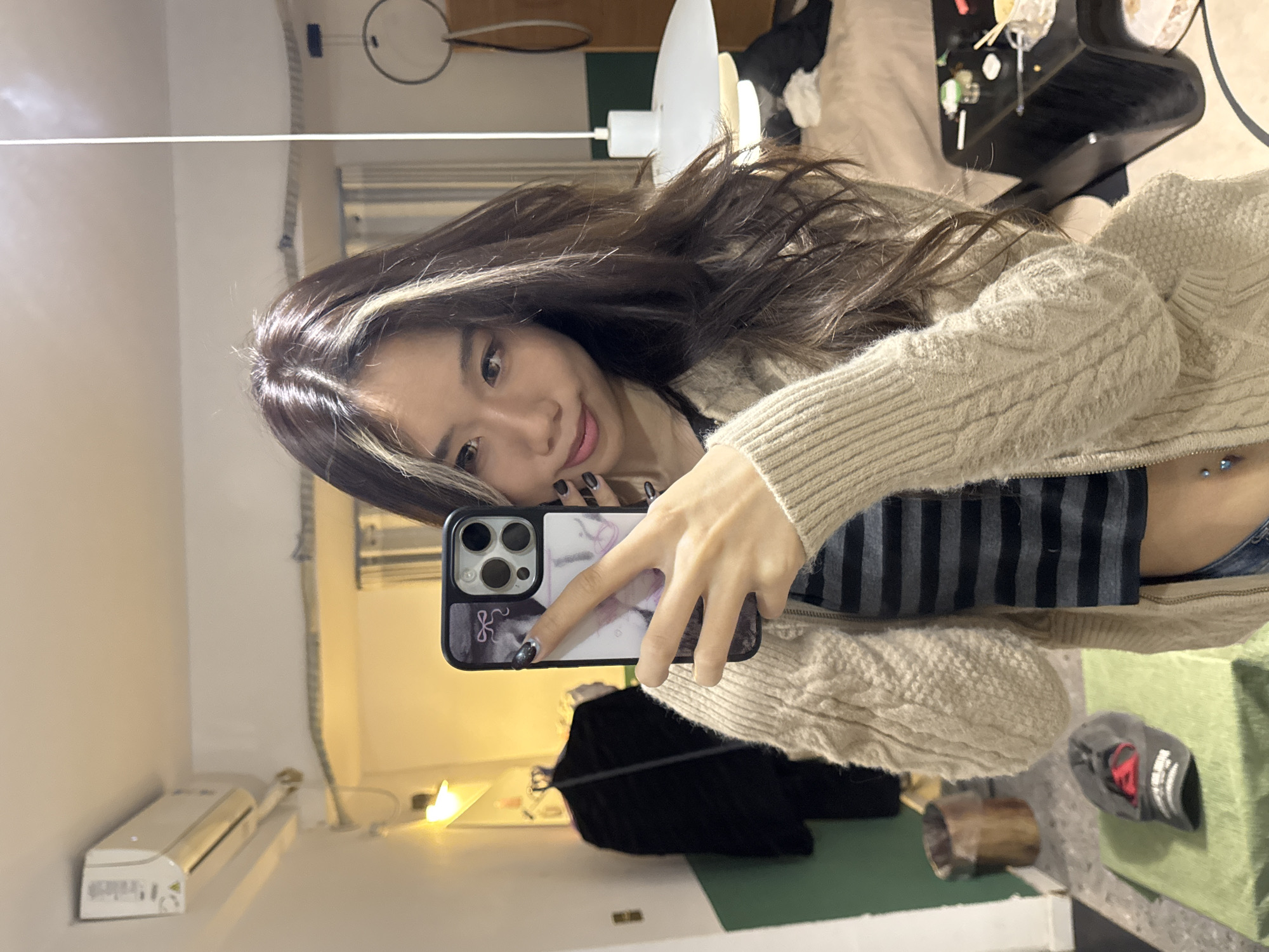 This image depicts an indoor scene in Liangjiang, China, captured as a mirror selfie. The central subject is a young East Asian female, shown from the chest up, with long dark brown hair featuring a prominent blonde streak on the viewer's left side, parted in the center. She has dark eyes, wears subtle makeup including pink lipstick, and displays a slight smile while looking towards the phone's lens. Her attire includes a beige cable-knit zippered cardigan worn over a black and grey striped crop top, exposing her midriff which features a navel piercing with a blue jewel. Her fingernails are painted dark grey. She holds a dark-colored smartphone, equipped with a multi-lens camera module, in a light-colored case adorned with a purple bow design, positioned to capture her reflection.
The reflected background reveals a domestic interior with white walls, a section of which has a green lower half. On the upper left, a white split-unit air conditioner is mounted. Below it, a warm-toned wall lamp illuminates a dark jacket hanging on a hanger. A dark-colored waste bin is on the floor to the left, adjacent to a green fabric-covered surface holding a grey baseball cap with a red logo. In the upper right, a white multi-tiered pendant lamp is suspended from the ceiling. A dark, geometrically shaped coffee table occupies the lower right, displaying remnants of a meal, including chopsticks, small dishes, and disposable containers. A bed with light-colored bedding is partially visible behind the table.