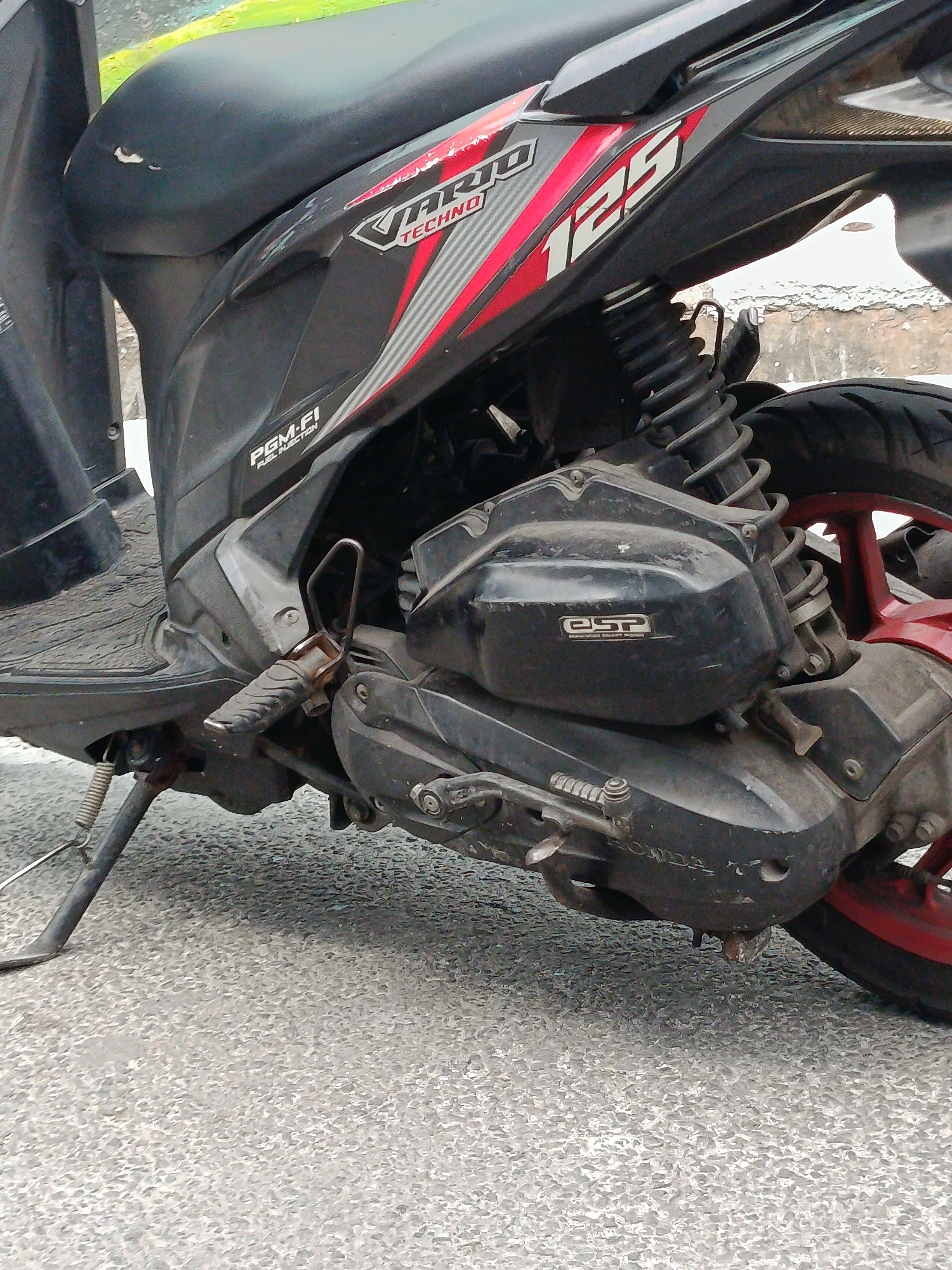 The image shows a close-up, low-angle view of the side and rear of a black and red scooter, specifically the Honda Vario Techno 125. The scooter is parked on an asphalt surface that appears to be a street or roadway. The scooter's engine casing, rear shock absorber, tire, and part of the seat are visible. The scooter has "PGM-FI FUEL INJECTION" and "ESP" logos on its body. The environment appears to be outdoors, with a blurry background that suggests a light-colored structure or wall behind the scooter. The scooter is dirty, indicating it has been used. The scene is devoid of people, and there are no discernible actions taking place. The lighting suggests it is daytime. The location is East Jakarta, Indonesia, as indicated by the provided context.