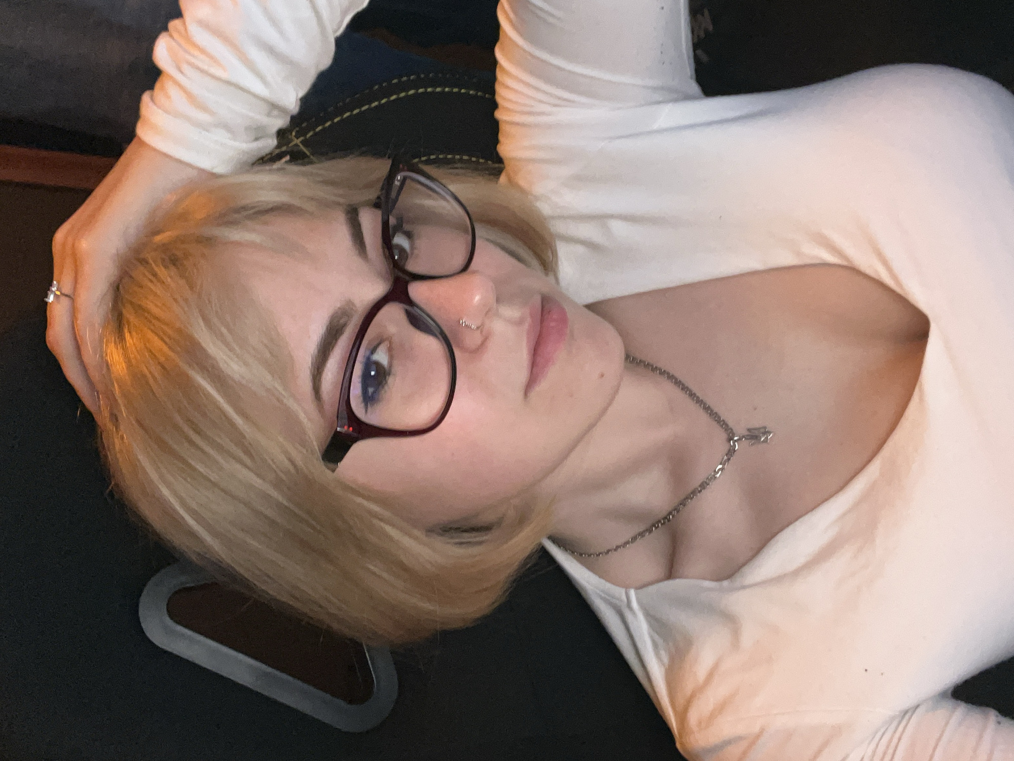 A young woman with blonde hair and fair skin is depicted in a close-up portrait, reclining and looking directly at the camera. She wears dark-framed eyeglasses, a small hoop nose piercing on her right nostril, and a silver ring on her left hand's ring finger. Her expression is neutral and engaging. She is dressed in a white long-sleeved top and a thin silver chain necklace with a small, indistinct pendant around her neck. Her left hand is raised, resting behind her head, prominently displaying the ring. The background is dark and indistinct, suggesting an indoor setting like a couch or bed. A dark object with a silver-grey trim is partially visible in the bottom left corner, possibly a mouse pad or part of a device. The lighting is soft and even, characteristic of indoor photography, likely in low-light conditions. No text is visible in the image. The location context provided is Budva, Montenegro.