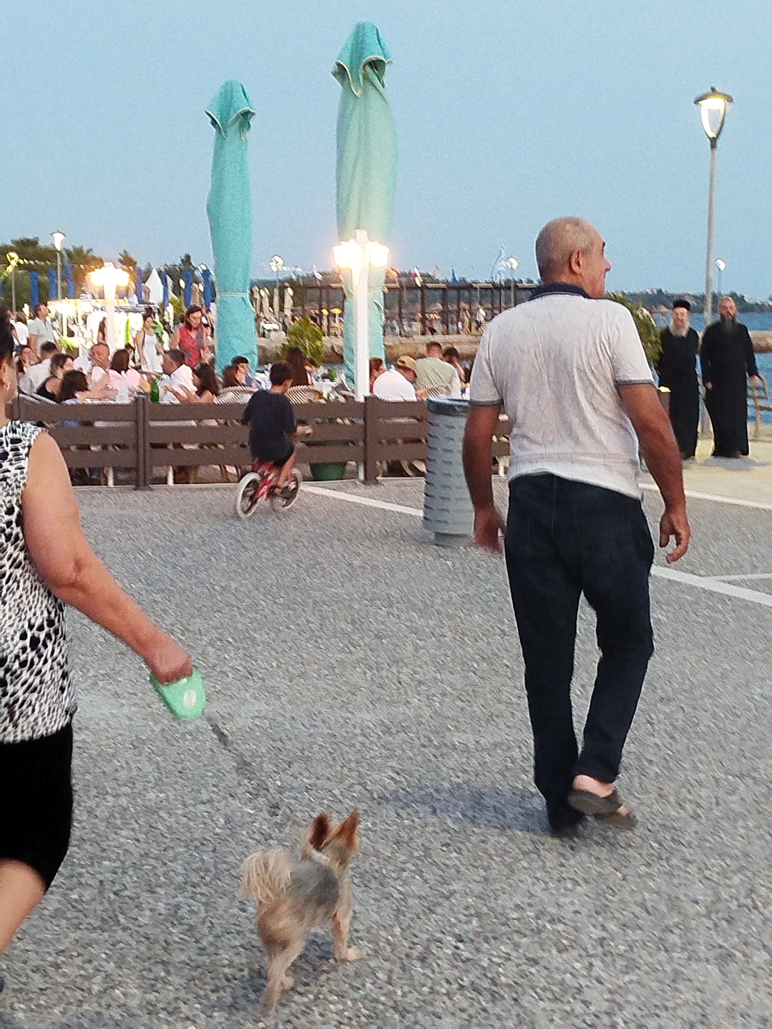 The image depicts an outdoor scene at dusk, possibly in the city of Sithonia Municipal Unit, Greece, characterized by a paved walkway and a body of water in the background. In the foreground, a woman on the left, wearing a black and white patterned top and a black skirt, is walking a small, light brown dog on a green leash. The dog is facing away from the camera. To the right of the woman, a bald man in a white t-shirt and dark pants is walking away from the camera, with his back partially turned. Further in the background, a young boy on a small red bicycle rides past a wooden fence. Beyond the fence, a seated group of people are visible at tables, suggesting an outdoor dining area. Large, light blue umbrella-like structures are prominently displayed throughout the background, some appearing to be closed. Two men in dark robes and head coverings are walking near a lamppost on the right side of the frame. The sky is a pale blue, indicating twilight.