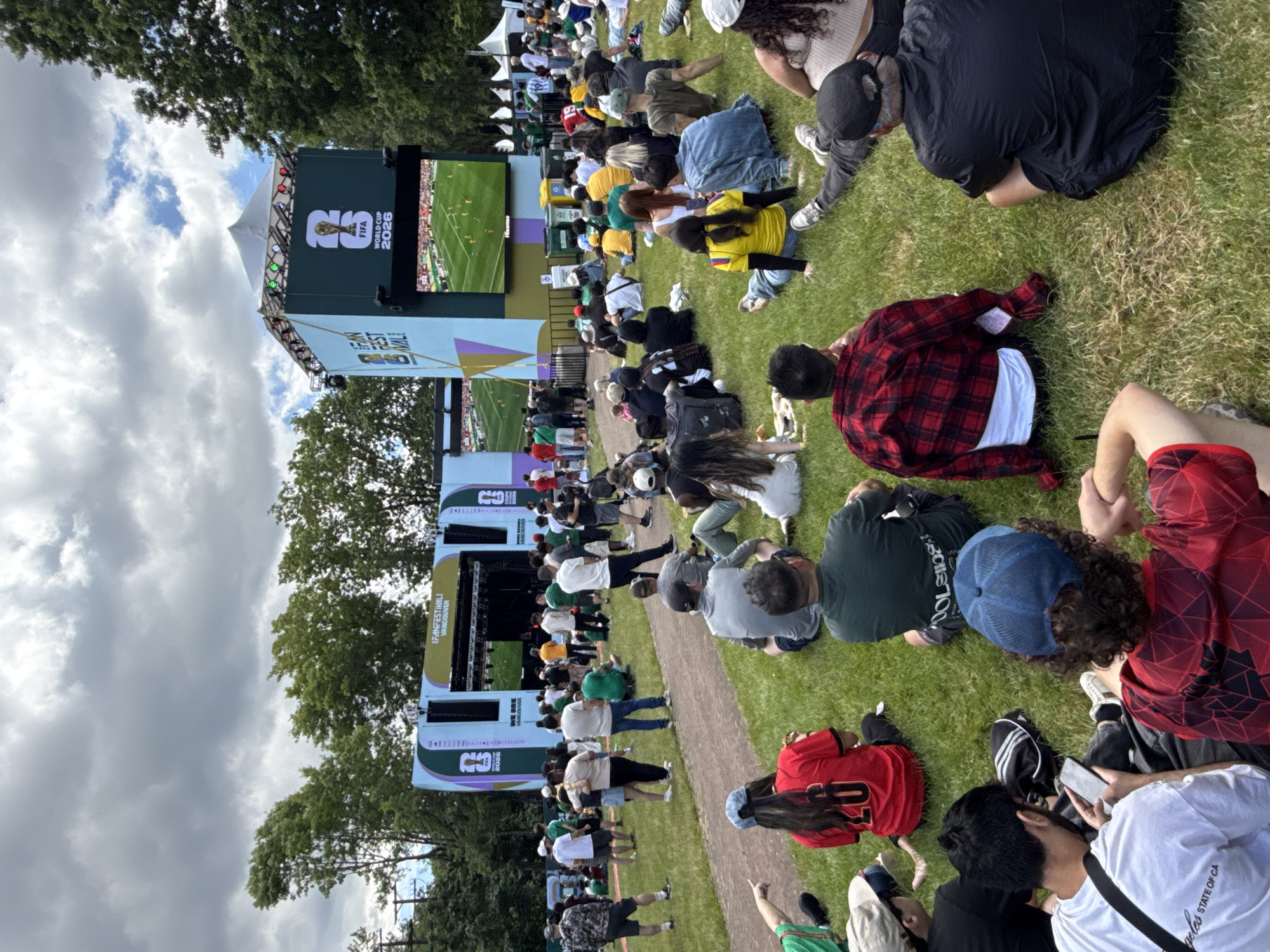 This image depicts a lively outdoor gathering at the FIFA Fan Festival Vancouver 2026, held in a grassy park area with trees in the background. A large screen displays a soccer match, drawing the attention of a diverse crowd seated on the grass. The atmosphere appears casual and festive, with many attendees dressed in casual attire, some wearing sports jerseys. The overall scene captures the excitement and communal experience of watching a major sporting event in an urban park setting.