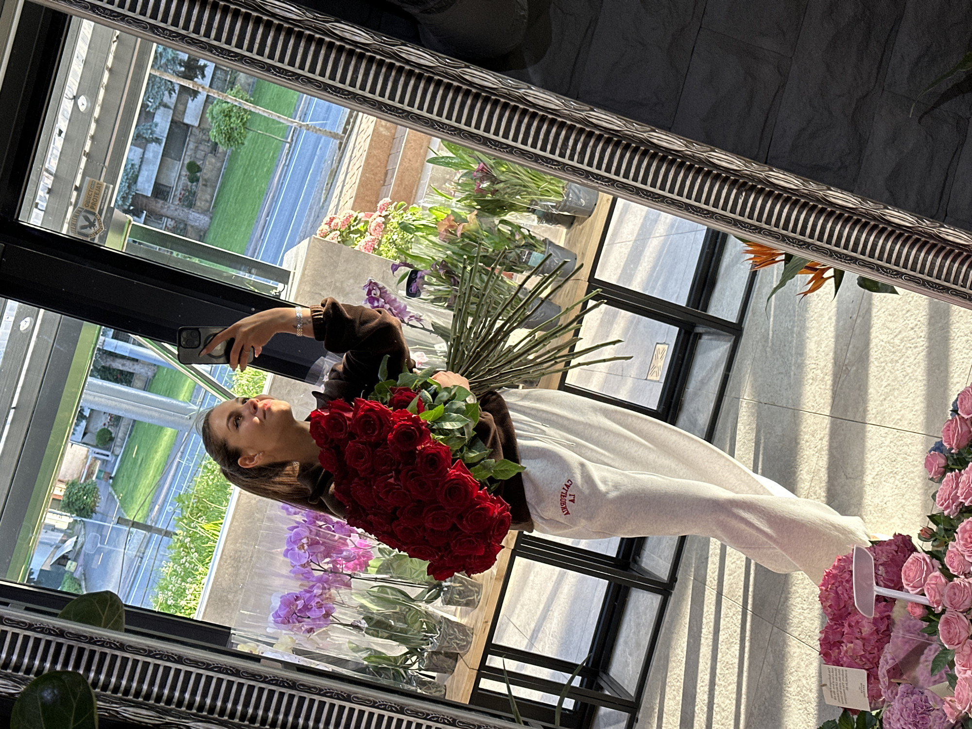 A young woman with her hair in a ponytail is reflected in a large, ornate mirror, taking a selfie with her phone. She is holding a large bouquet of red roses and wearing a brown sweatshirt and light-colored sweatpants with red text on the left leg. In the background, visible through a window and also reflected in the mirror, is a street scene with a grassy area, trees, and buildings. Various potted orchids and other plants are displayed on shelves behind the woman. The overall setting appears to be a flower shop or a similar commercial establishment in Chișinău, Moldova.