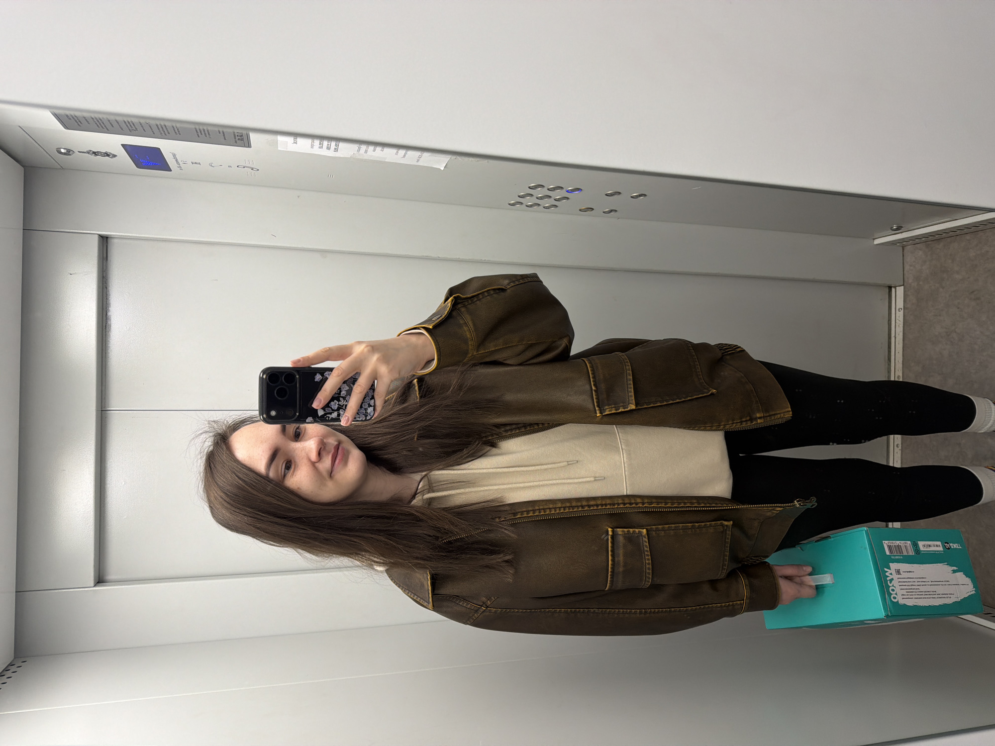 A young woman is taking a selfie in an elevator. She is wearing a brown jacket, a light-colored hoodie, and black leggings. She is holding a turquoise box in her left hand. The elevator has white walls and a control panel with buttons and an indicator display. The location is likely in the city of Chaikovsky, Russia, as indicated on the box she is holding.