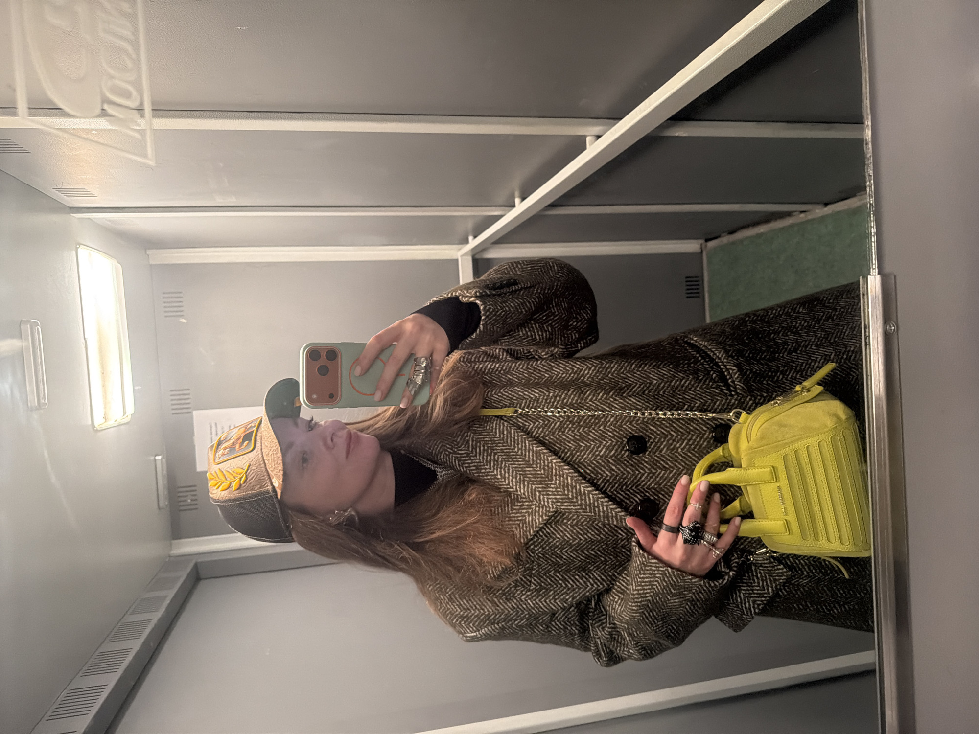 The image depicts a woman taking a selfie in what appears to be an elevator or a small room with mirrored walls. The walls are light gray. She is wearing a brown herringbone coat, a black turtleneck, and a baseball cap with gold embellishments. She has long brown hair. She is holding a smartphone with a light green case to take the photo. She is also wearing several rings on her fingers. A small yellow purse is slung across her body. The background details are limited, but there are some vents visible on the wall and a light fixture on the ceiling. There is no indication of Moscow in this image.