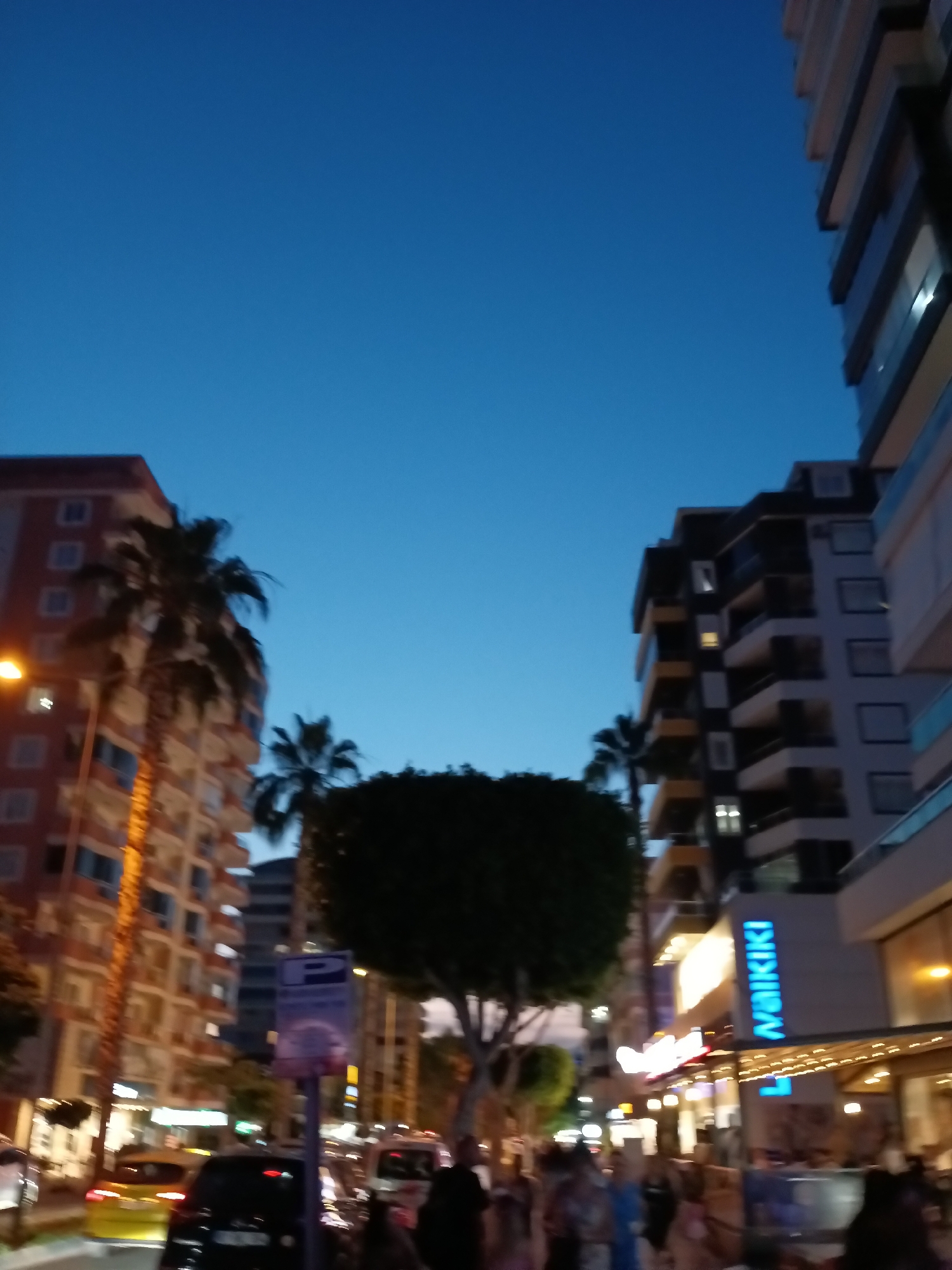 The image depicts a street scene in Alanya, Turkey, during twilight hours. Tall residential or hotel buildings with balconies line both sides of the street, interspersed with palm trees. A bright blue sign with the text "WAIKIKI" in vertical, illuminated lettering is visible on the right side of the image, indicating a commercial establishment. Pedestrians are present on the sidewalks, some appearing to be gathered around shopfronts and cafes. Vehicles, including a yellow car and a black car, are parked or driving on the street. The overall atmosphere suggests a bustling urban environment as evening descends, with artificial lighting beginning to illuminate the scene. The sky is a clear, deep blue, indicative of dusk.
