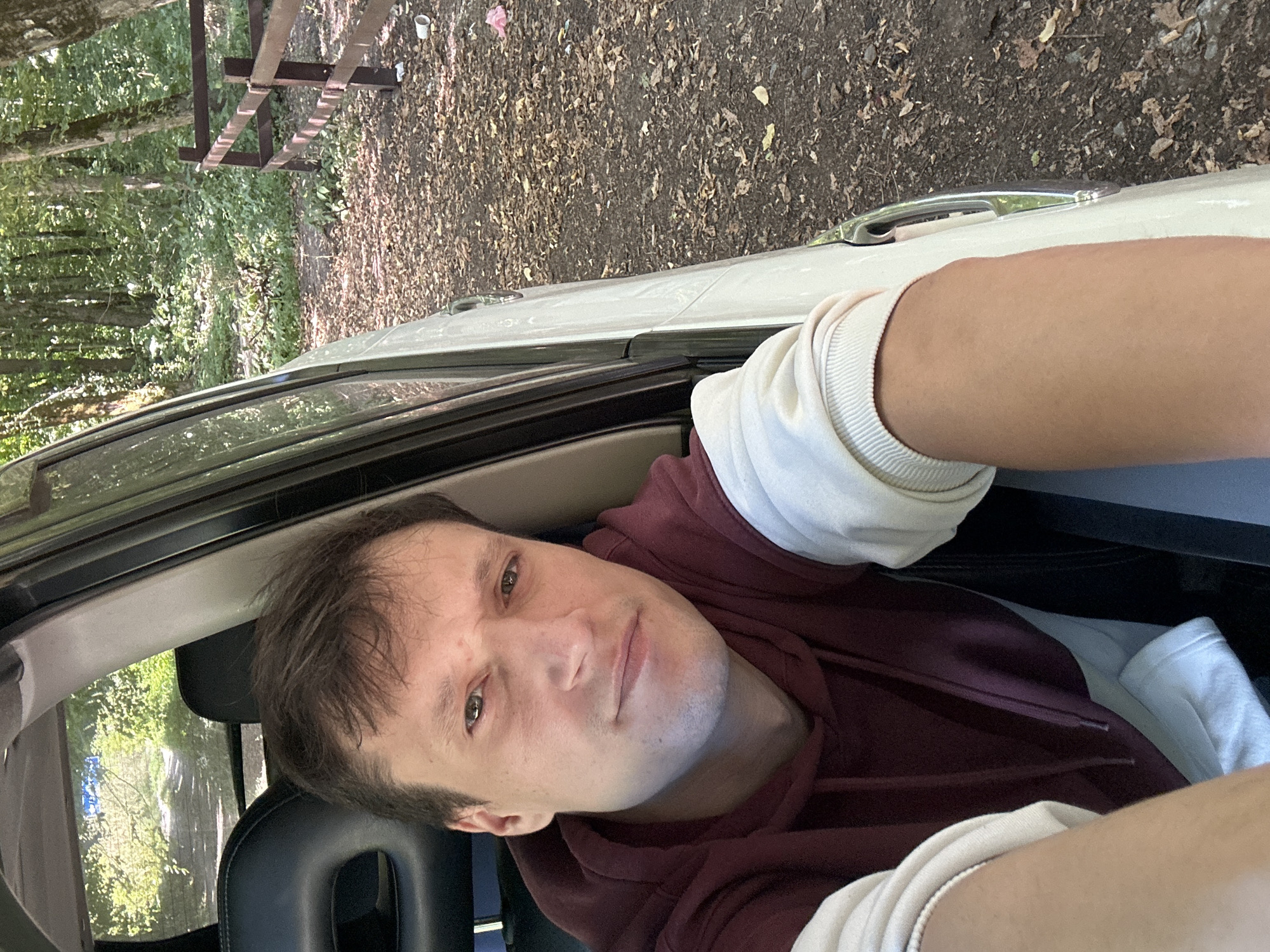 A man is sitting in the driver's seat of a car, looking at the camera. The car is parked on a dirt road surrounded by trees and foliage. A wooden fence is visible to the right of the car. The ground is covered with fallen leaves and some litter. The man is wearing a burgundy hooded sweatshirt with cream-colored sleeves and a white t-shirt underneath. The lighting suggests it is daytime, with sunlight filtering through the trees.