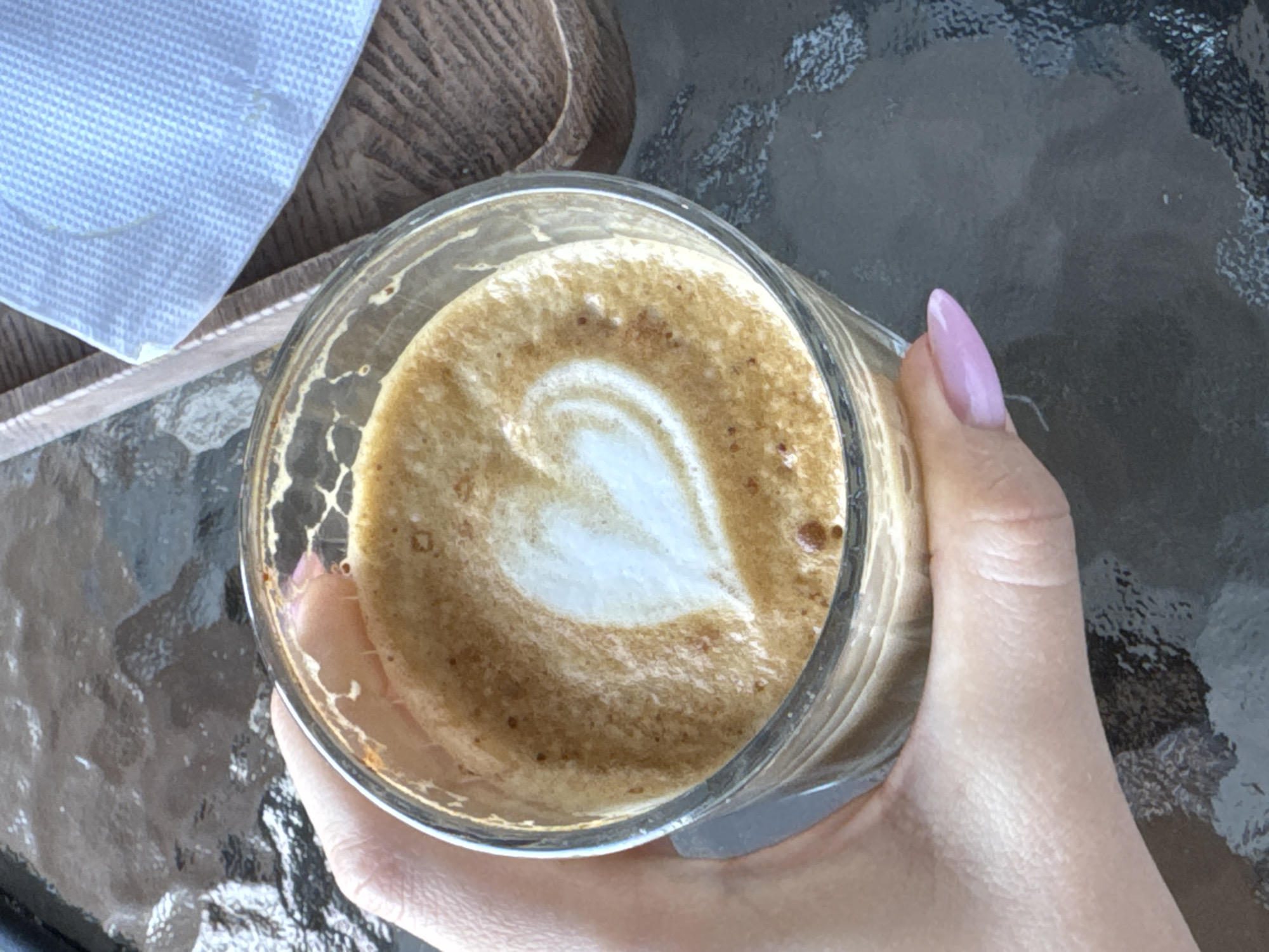 A close-up shot shows a hand holding a clear glass of coffee with latte art in the shape of a heart. The coffee is a light brown color with a creamy white foam on top. The background is a dark, textured surface, possibly a table. To the left, a portion of a light blue napkin is visible, placed on a wooden tray. The hand holding the coffee has a light pink nail polish. The lighting suggests it might be daytime, possibly outdoors given the textured table. There is no visible text in the image.