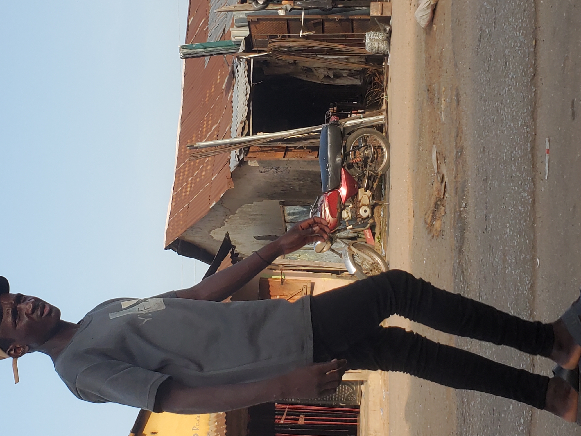 A street scene in Mararaba, Nigeria, features a dark-skinned man in a gray t-shirt and black pants, standing on a paved road. He faces left with his right arm extended, appearing to hold something. Behind him is a weathered structure with a corrugated metal roof and a dark opening filled with metal objects. A motorcycle is partially visible inside the structure. The street is narrow with signs of wear and debris. The sky is visible in the upper portion of the frame.
