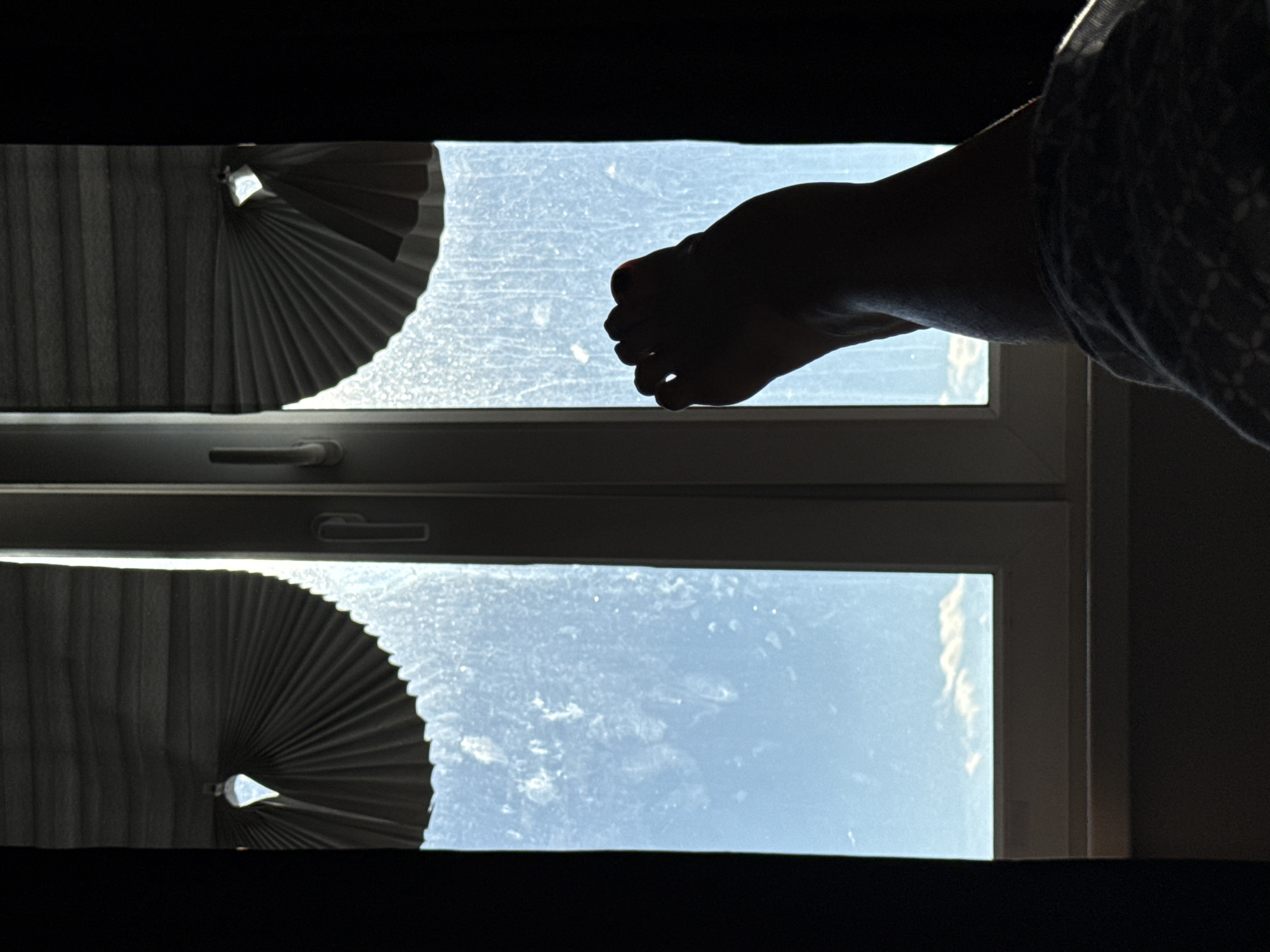 A silhouette of a person's foot and lower leg is visible in the foreground of the image, positioned in front of a window. The foot is dark, with distinct toes, and is resting on a patterned blanket. The window is divided into two panes, with a white frame. The left pane of the window is partially covered by a pleated blind that is fan-shaped and angled downwards. The right pane of the window is mostly open, revealing a bright blue sky with some scattered clouds. The window itself appears slightly dusty or smudged. Dark curtains frame the sides of the window, adding to the contrast between the illuminated sky and the shadowed interior. The scene suggests a moment of relaxation or observation from indoors.