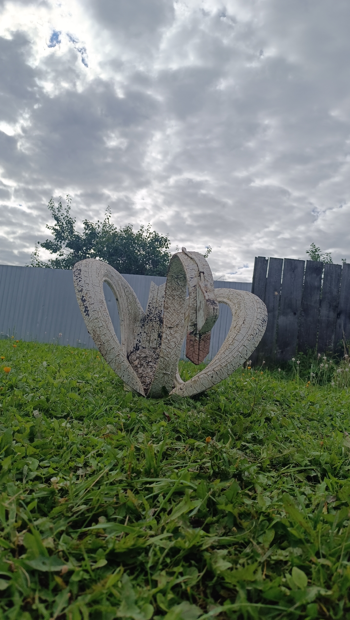 The image depicts a white, weathered sculpture made from what appears to be old tires, set on a patch of green grass in an outdoor environment. The sculpture has a vaguely heart-like or floral shape, with segments of tire forming curved petals or lobes. The material is cracked and peeling, showing signs of age and exposure to the elements. In the background, a grey metal fence runs horizontally across the frame, partially obscured by trees. To the right of the sculpture, a weathered wooden fence stands at an angle. The sky above is overcast with scattered clouds. The scene is set in a yard or open area, with no people or animals visible. The location is identified as Karsha, Russia.
