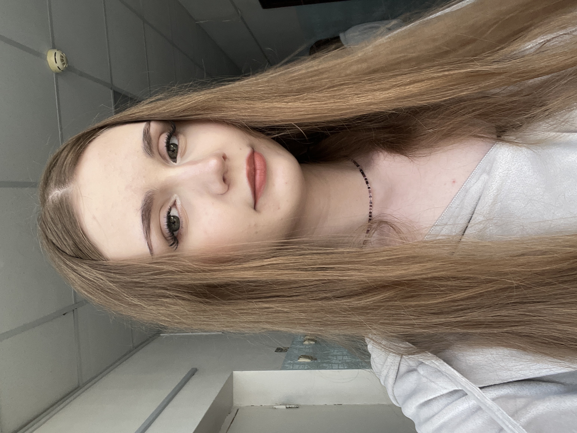 A young woman with long, straight, light brown hair is captured in a selfie. She has light skin, green eyes, and is wearing subtle makeup, including eyeshadow and lipstick. Around her neck, she wears a beaded necklace. She is dressed in a light gray, zip-up hoodie.
The background suggests an indoor environment, possibly a hallway or corridor. The ceiling appears to be a suspended panel ceiling, common in institutional or public buildings. A smoke detector is visible on the ceiling. To the left of the woman, a white door and part of a wall with some blue tiling are visible. To the right, a dark doorway or opening is present, with a blurry figure of another person visible in the background, possibly also indoors.
While the exact location within Sterlitamak, Russia, cannot be determined from the image alone, the indoor setting with features like suspended ceilings and tiled walls could indicate a school, hospital, office building, or similar public facility. The lighting appears to be artificial, typical of indoor environments.
