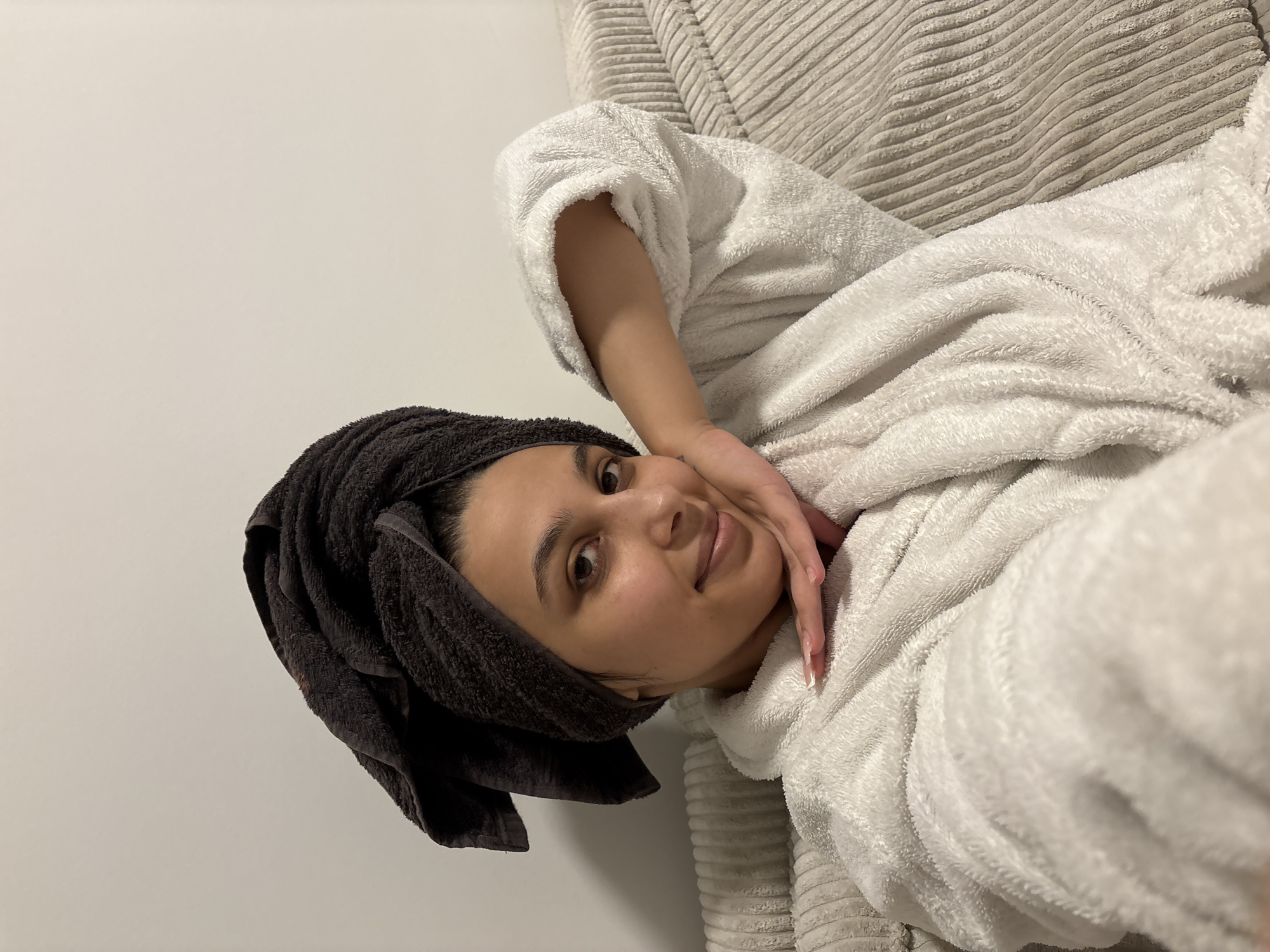 A young woman is depicted in a close-up, horizontal portrait, reclining in what appears to be an indoor setting. She is wearing a soft, white, fluffy bathrobe and has a dark grey or black towel wrapped around her head, suggesting a post-shower or relaxed state. Her head is tilted slightly, and she looks directly at the camera with a gentle, relaxed smile. Her left hand is casually placed near her chin and jawline, showcasing long, manicured nails with light-colored tips.
The background behind her features a plain, light-colored wall on the left side, and a textured, light grey ribbed material on the right, which could be a cushion, pillow, or part of a couch or bed. The overall lighting is soft and even, highlighting the textures of her attire and the surroundings. There is no visible text in the image. The scene conveys a sense of comfort and tranquility, possibly during a leisure moment in a home in Mönchengladbach, Germany.