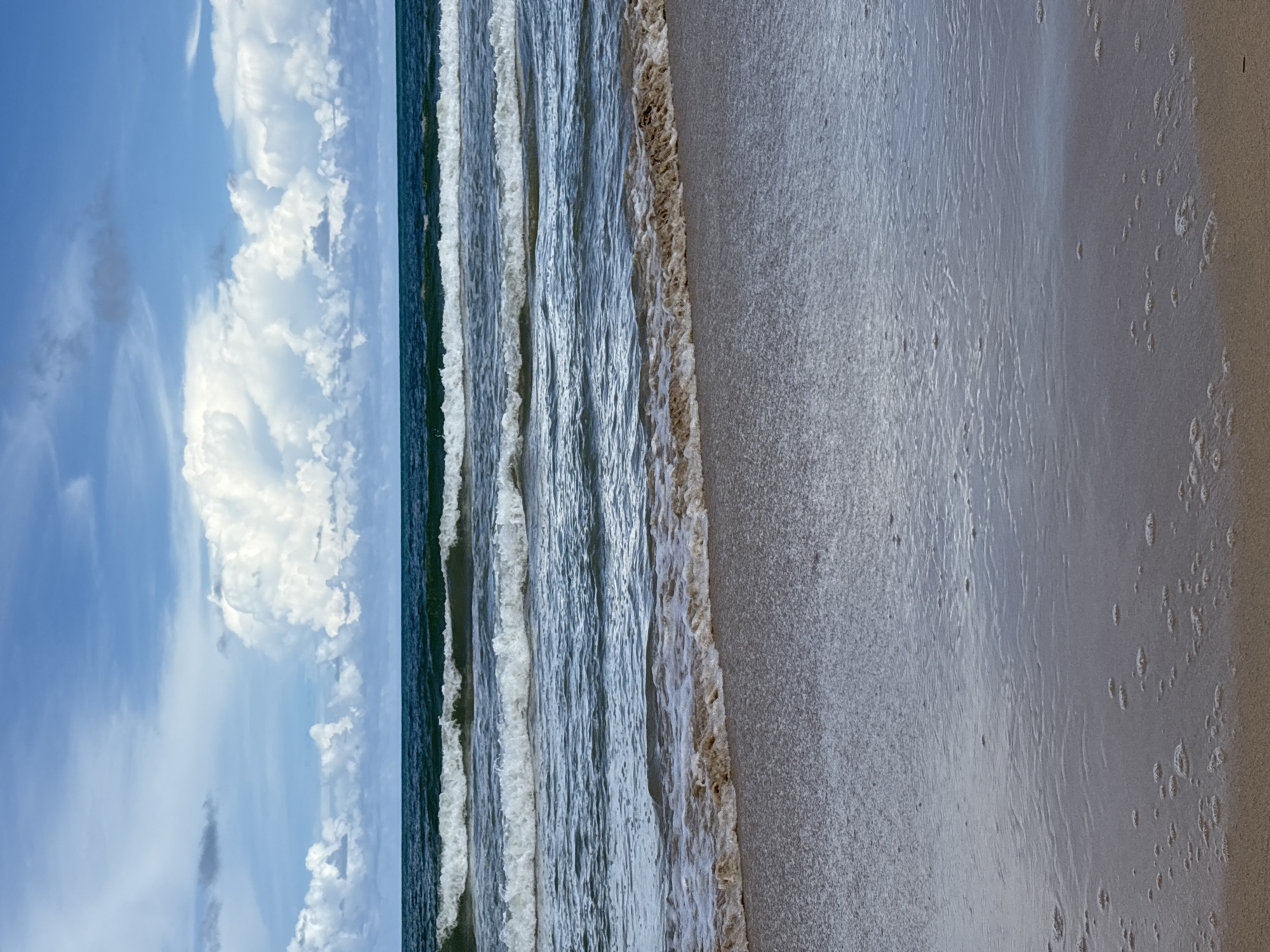 The image depicts a tranquil beach scene with the ocean stretching towards the horizon under a partly cloudy sky. The water is a deep blue, with several layers of white-capped waves rolling towards the shore. The sand in the foreground is wet and reflects the light, with small bubbles scattered on its surface. The clouds in the sky are dramatic and voluminous, with some casting shadows on the water. The scene appears to be a natural environment, likely a public beach within the city of Choeng Thale, Thailand, given the tropical appearance of the clouds and the overall ambiance. There are no people or man-made objects visible in the image, focusing the viewer's attention entirely on the natural elements of the beach, sea, and sky.