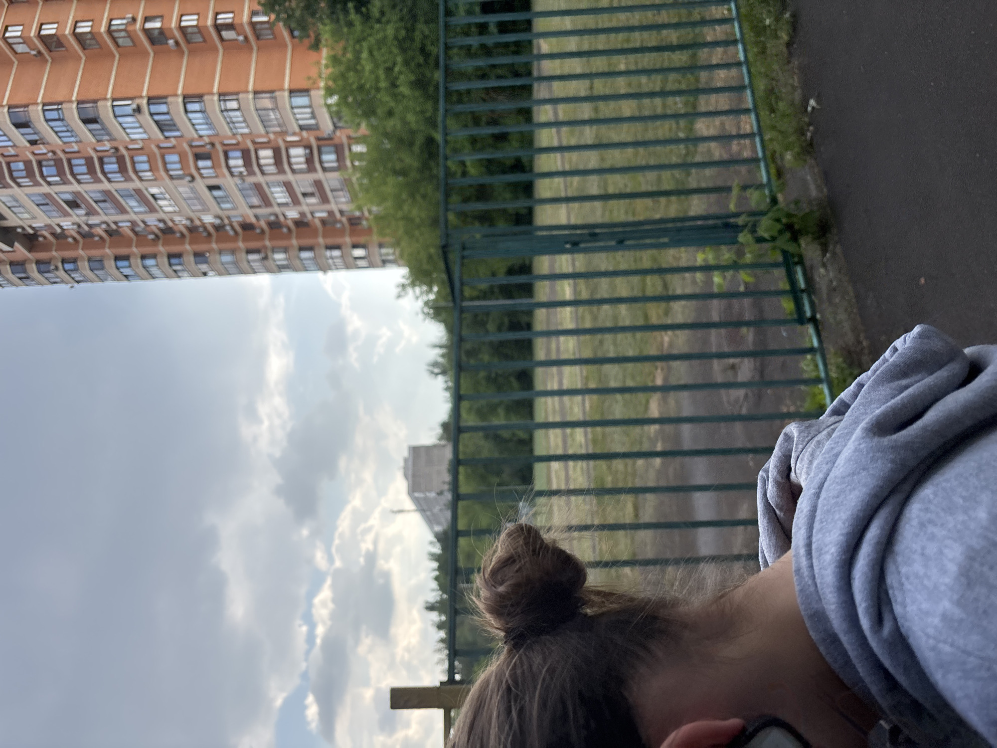 An outdoor medium shot captures a young person from behind, their head and shoulders visible, looking out at a fenced-off grassy area. The person has their dark hair pulled into a bun. Behind them, a tall, orange brick apartment building with many windows stands against a cloudy sky. Further in the background, a long, light-colored building can be seen. A green metal fence runs horizontally across the mid-ground, separating the viewer from the grassy field and the distant structures. The foreground shows a dark asphalt surface, likely a path or road. The scene is set in an urban environment, likely a residential area in Moscow, Russia, given the architecture of the buildings and the style of the apartment complex.