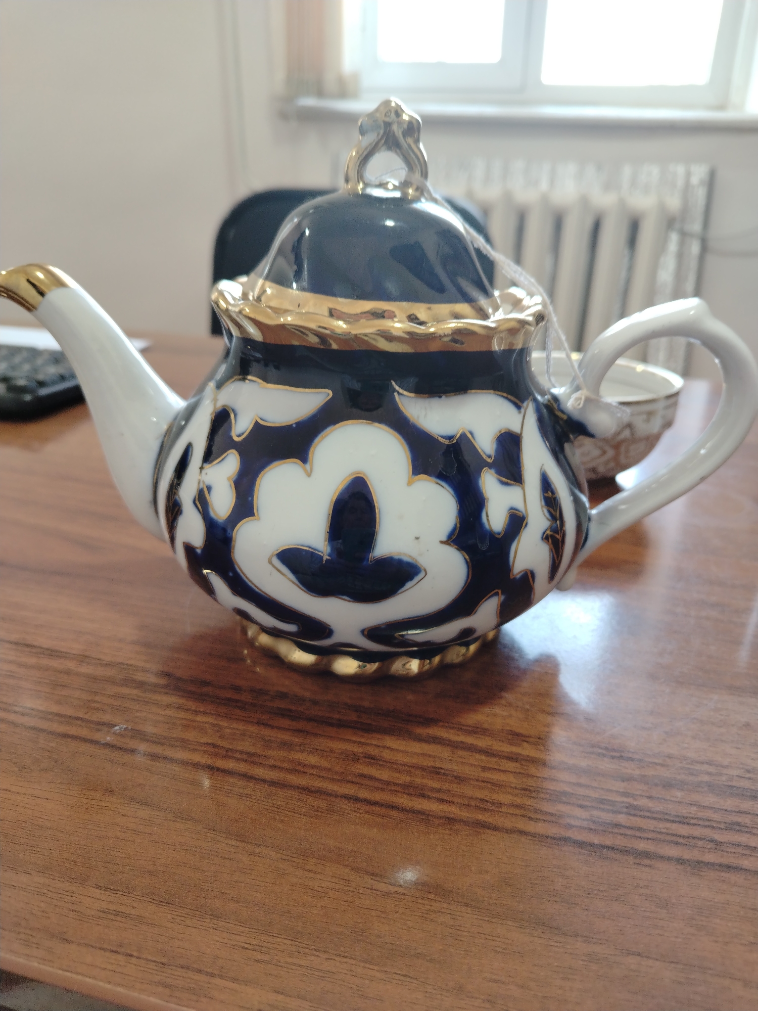 A close-up, eye-level shot captures a white ceramic teapot with a dark blue floral pattern and gold trim. The teapot is positioned on a polished wooden table, reflecting the light. In the background, a radiator and a window are visible, suggesting an indoor setting, possibly in Nurata, Uzbekistan, given the context. The teapot has a curved handle and a spout, and its lid is partially covered in plastic. A white string is attached to the lid, possibly for identification or decoration. The overall impression is one of domesticity or a shop display.
