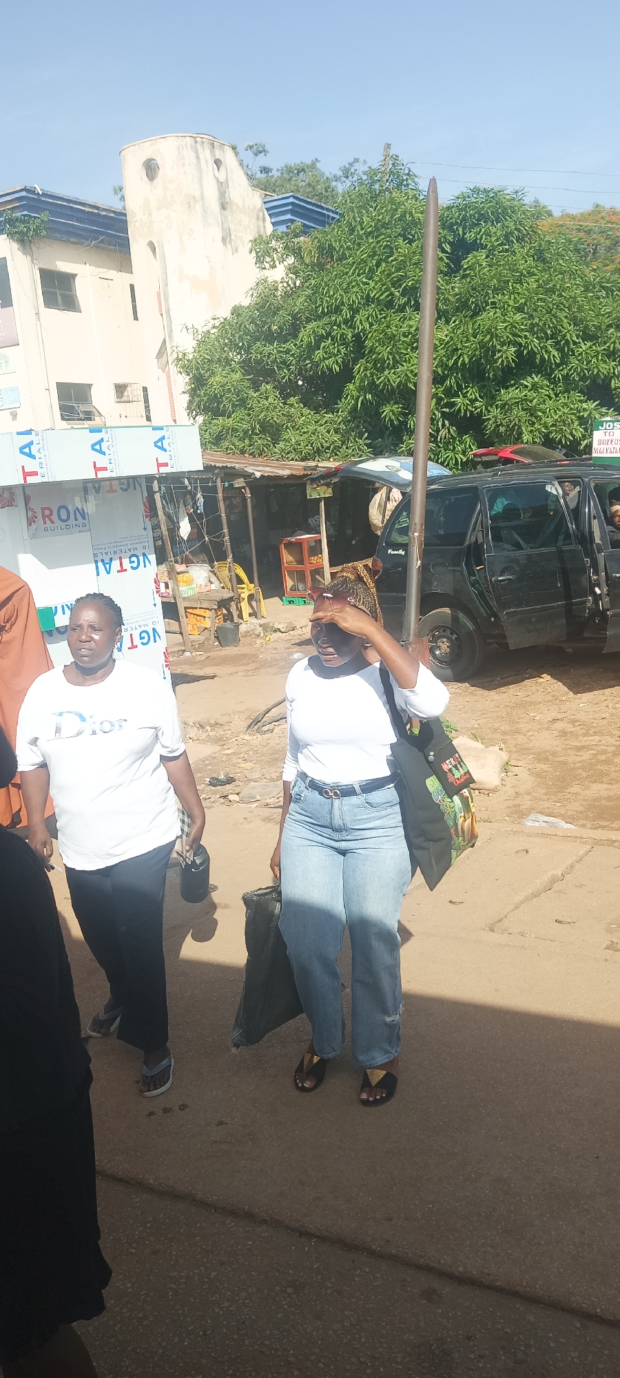 This outdoor image captures a sunny street scene, likely a market or commercial area in Jos, Nigeria. In the foreground, two women are walking. The woman on the left wears a white t-shirt with "Dior" printed on it, black pants, and sandals, carrying a black thermos or flask. The woman on the right is dressed in a white long-sleeved top, blue jeans, and black sandals with gold accents. She carries a black tote bag, which appears to have "MERRY" and "LAND" text. She shields her eyes with her hand, indicating bright sunlight, and has sunglasses perched on her head. A third person is partially visible on the far left.
The background reveals typical urban infrastructure, including market stalls with corrugated metal roofs and wooden frameworks. Various signs for building materials, such as "TAI," "RON BUILDING," and "NGTAI MATERIALS," are visible on a nearby structure. A black SUV or minivan is parked on the right side of the street, with the word "Family" faintly visible on its side. A tall, thin metal pole stands prominently in the mid-ground.
Further in the background, a sign partially reads "JOS TO BOZKO MAIKATA," confirming the location in Jos, Nigeria, and possibly indicating a transport route or stop. Green trees and foliage are scattered throughout the scene. A light-colored building with a distinctive round, tower-like section featuring small circular windows is visible in the upper left, beneath a clear blue sky. The ground is a mix of dirt and cracked pavement, illuminated by bright daylight, suggesting mid-morning or early afternoon.