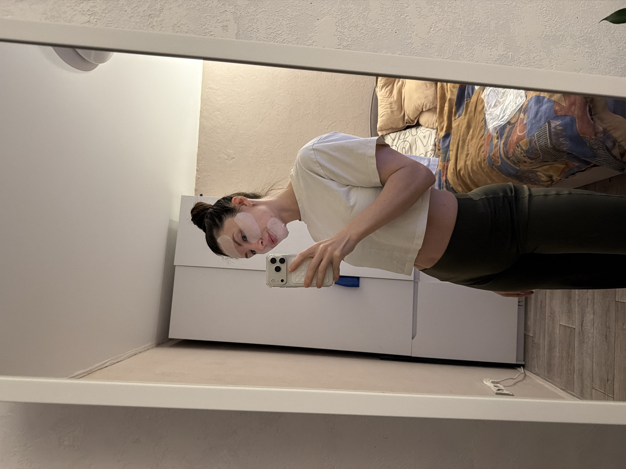 A young adult woman with bandages on her face is taking a selfie in a mirror. She is wearing a white t-shirt and dark green leggings. She is holding a white iPhone in her right hand. The background shows a bedroom with a bed, bedding, a white cabinet, and a wall. The location is a residence, likely in Perm, Russia.