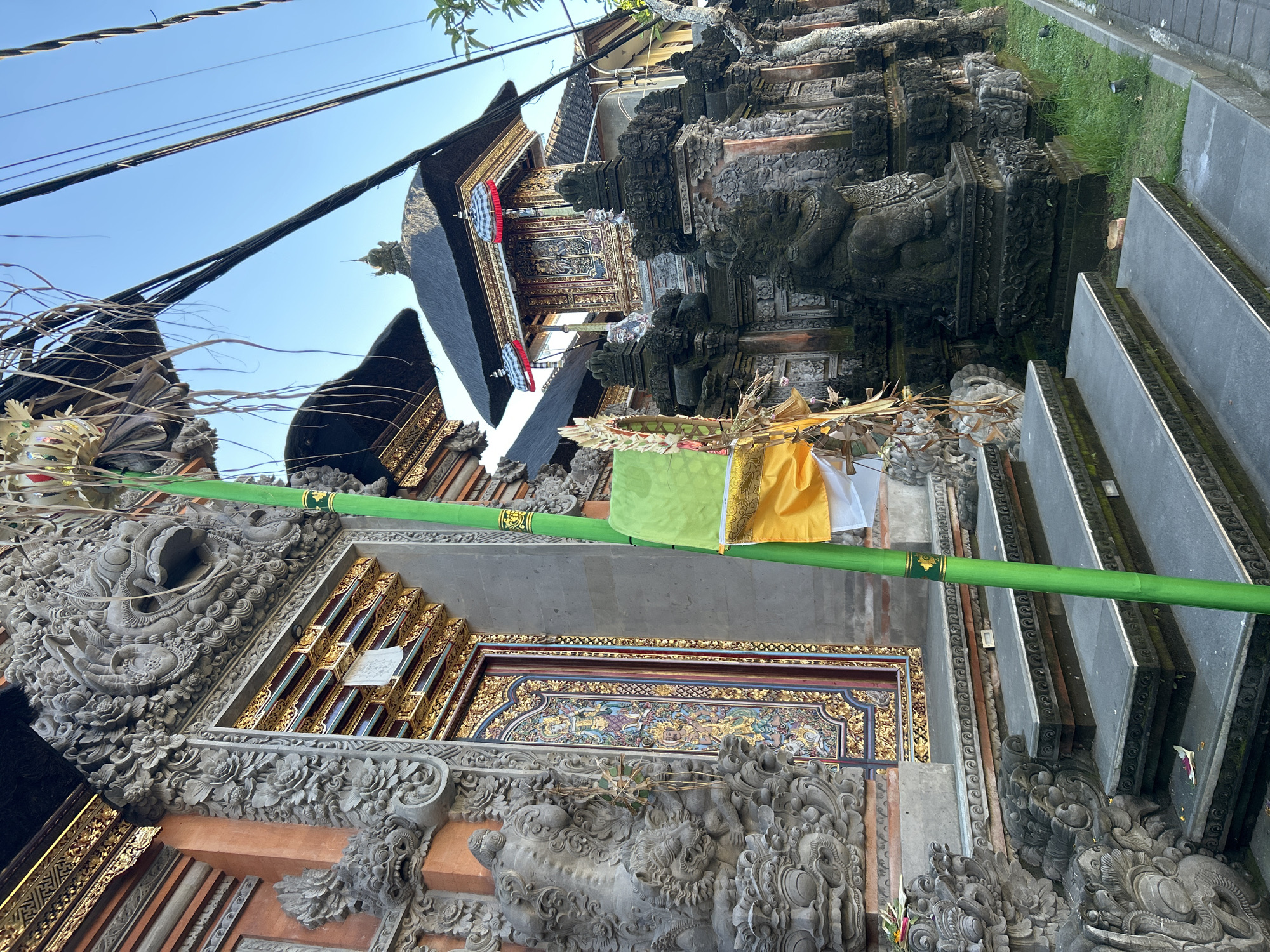 The image shows a close-up view of the entrance to a Balinese temple, likely in the Ubud District of Indonesia. The temple features intricate stone carvings on its facade, including decorative figures and a large, menacing face above the doorway. The door itself is ornately decorated with colorful paintings and gold accents.
A tall, green bamboo pole stands in front of the entrance, adorned with a yellow and green fabric banner and dried palm leaves, suggesting a religious or ceremonial offering. Steps lead up to the temple entrance, and stone statues of mythological creatures line the pathway.
In the background, another section of the temple with a thatched roof and golden ornamentation is visible. Overhead, a network of electrical wires cuts across the clear blue sky, indicating the proximity of a residential or commercial area. The overall scene depicts a blend of traditional Balinese architecture and contemporary infrastructure.