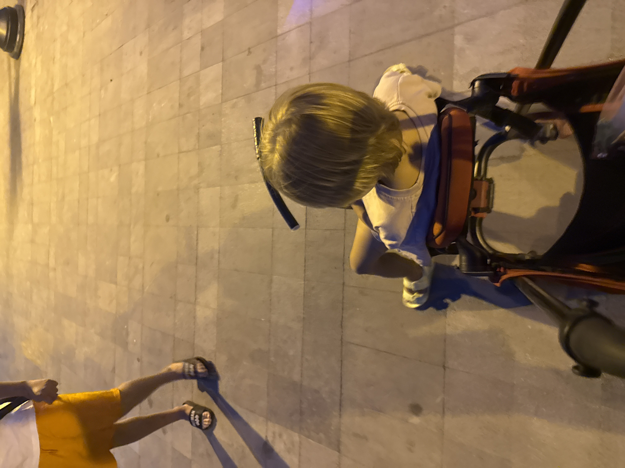 A child with blond hair is seated in a stroller, viewed from behind. The child is wearing a light-colored shirt. An adult, wearing orange shorts and white sandals, stands to the left of the frame, casting a shadow on the tiled ground. The scene appears to be an outdoor pedestrian area, likely a promenade or sidewalk, illuminated by artificial light, suggesting it is evening or nighttime. The location is identified as Rafailovići, Montenegro.