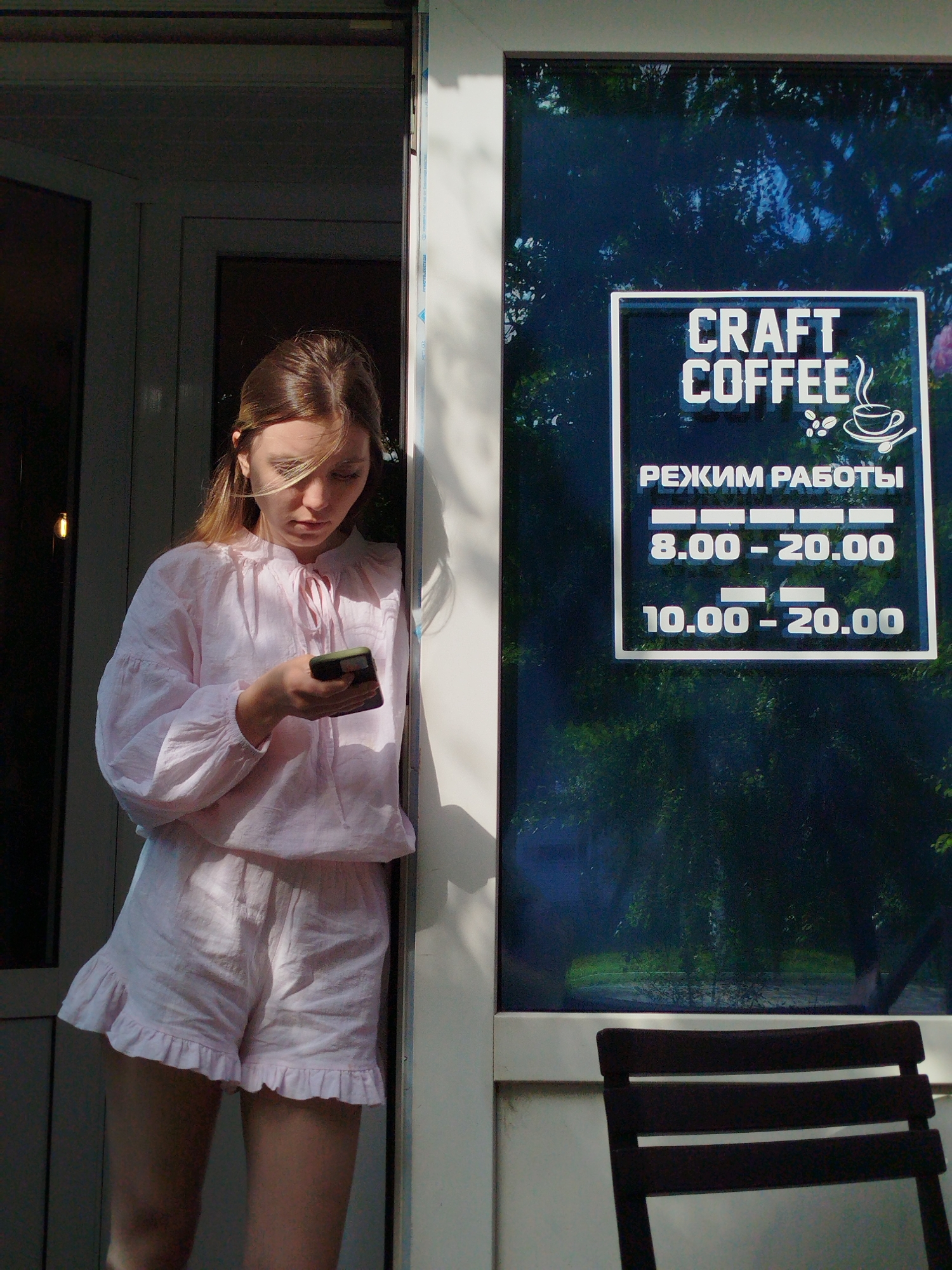 A young woman with blonde hair stands in a doorway, looking at her phone. She is wearing a pink blouse and shorts. To her right, a glass window displays a sign for "CRAFT COFFEE" with operating hours listed in Russian. The background outside the window appears to be a natural setting with trees. A dark chair is visible in the foreground of the window.