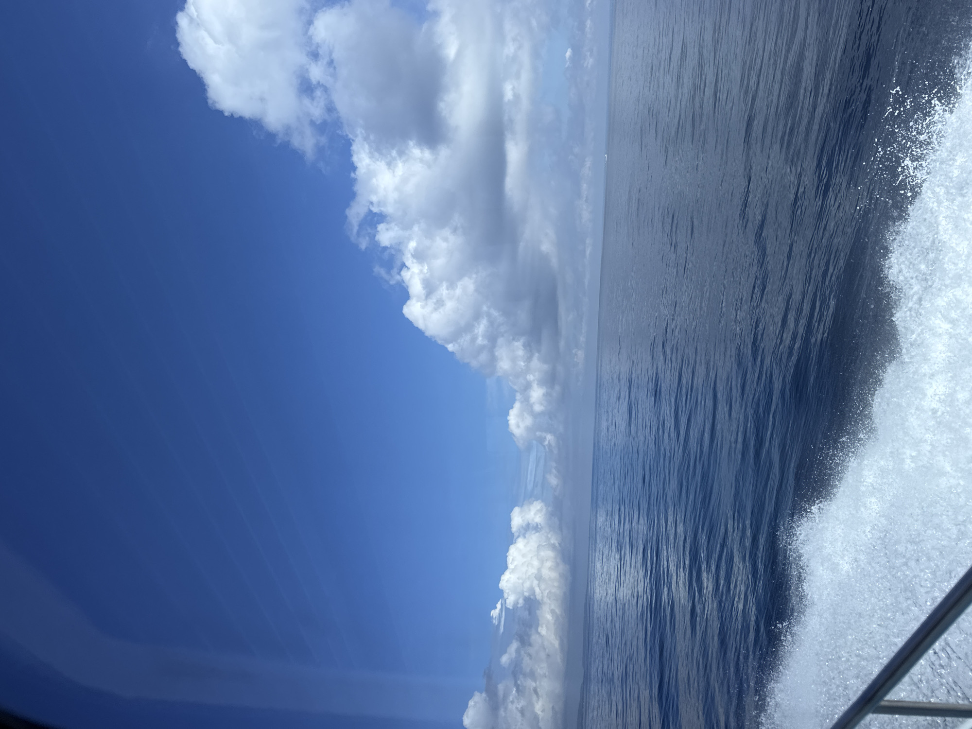 The image shows a view from a boat on the water. The water is dark blue and choppy, with white spray visible near the edge of the frame. The sky is bright blue with white clouds. Part of the boat's railing is visible in the lower right corner. Location is Unknown, Indonesia.