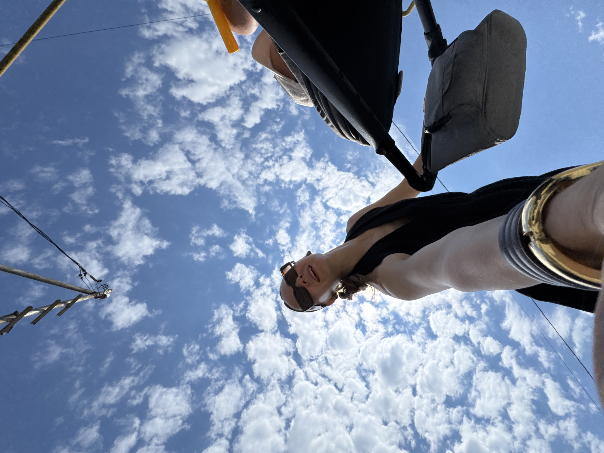 The image depicts a low-angle shot of a woman holding a baby stroller against a bright blue sky with scattered white clouds. The woman is wearing sunglasses and a black sleeveless dress, and a thick gold bangle is visible on her wrist. She is looking upwards and to her left, with her mouth slightly open as if speaking. Part of the baby stroller, including a dark fabric bag and a yellow handle, is visible. Overhead, a section of a utility pole with wires is visible against the sky. The scene appears to be outdoors, possibly in a park or on a street, under sunny weather conditions. The image does not contain any specific visual cues that would allow for the determination of the location being Sochi, Russia.