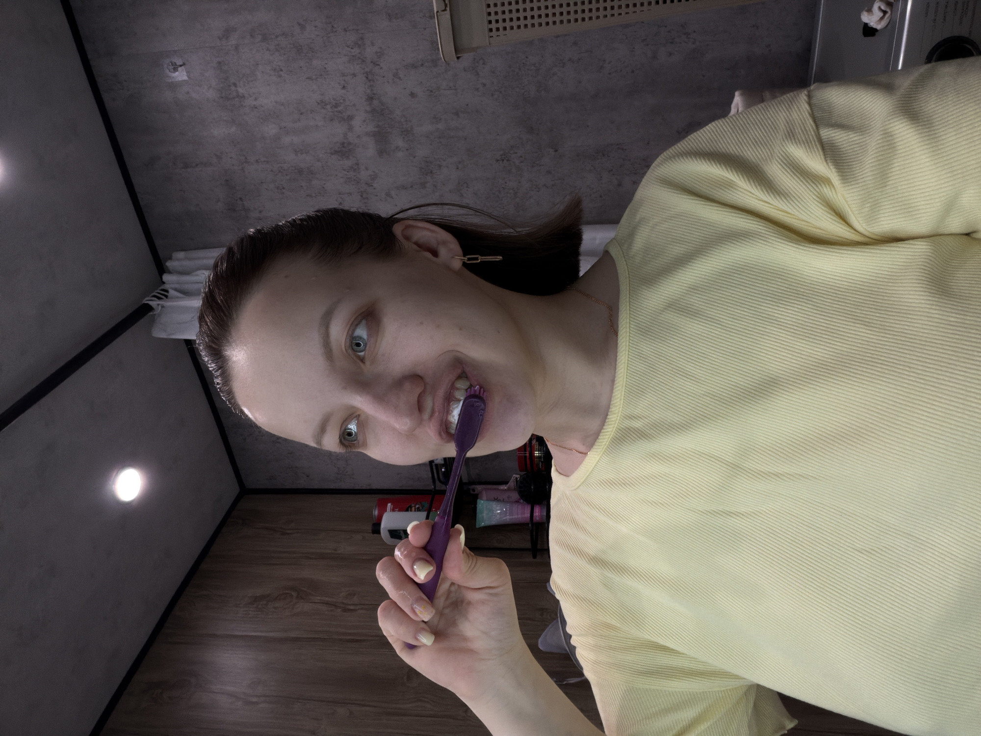 A woman with fair skin and light eyes is brushing her teeth. She is wearing a light yellow shirt and a gold chain necklace. Her hair is pulled back. She holds a purple toothbrush with white toothpaste in her right hand. The background appears to be a bathroom, with gray walls and a light fixture on the ceiling. Various personal care items are visible on a shelf behind her. There is no specific information suggesting the location is Reshetnikovo, Russia.
