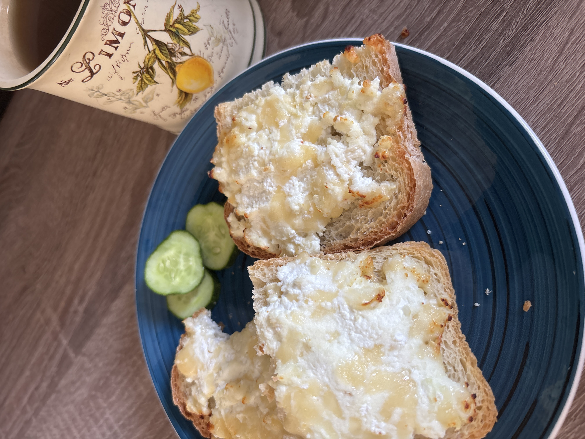 Two slices of toasted bread, topped with a white spread that appears to be cottage cheese or ricotta, sit on a dark blue plate. Next to the toast are three thin slices of cucumber. In the background, a mug with a lemon and script design is partially visible. The setting appears to be a home or cafe, with a wooden table surface. There is no information available about the specific location in Naberezhnye Chelny, Russia.