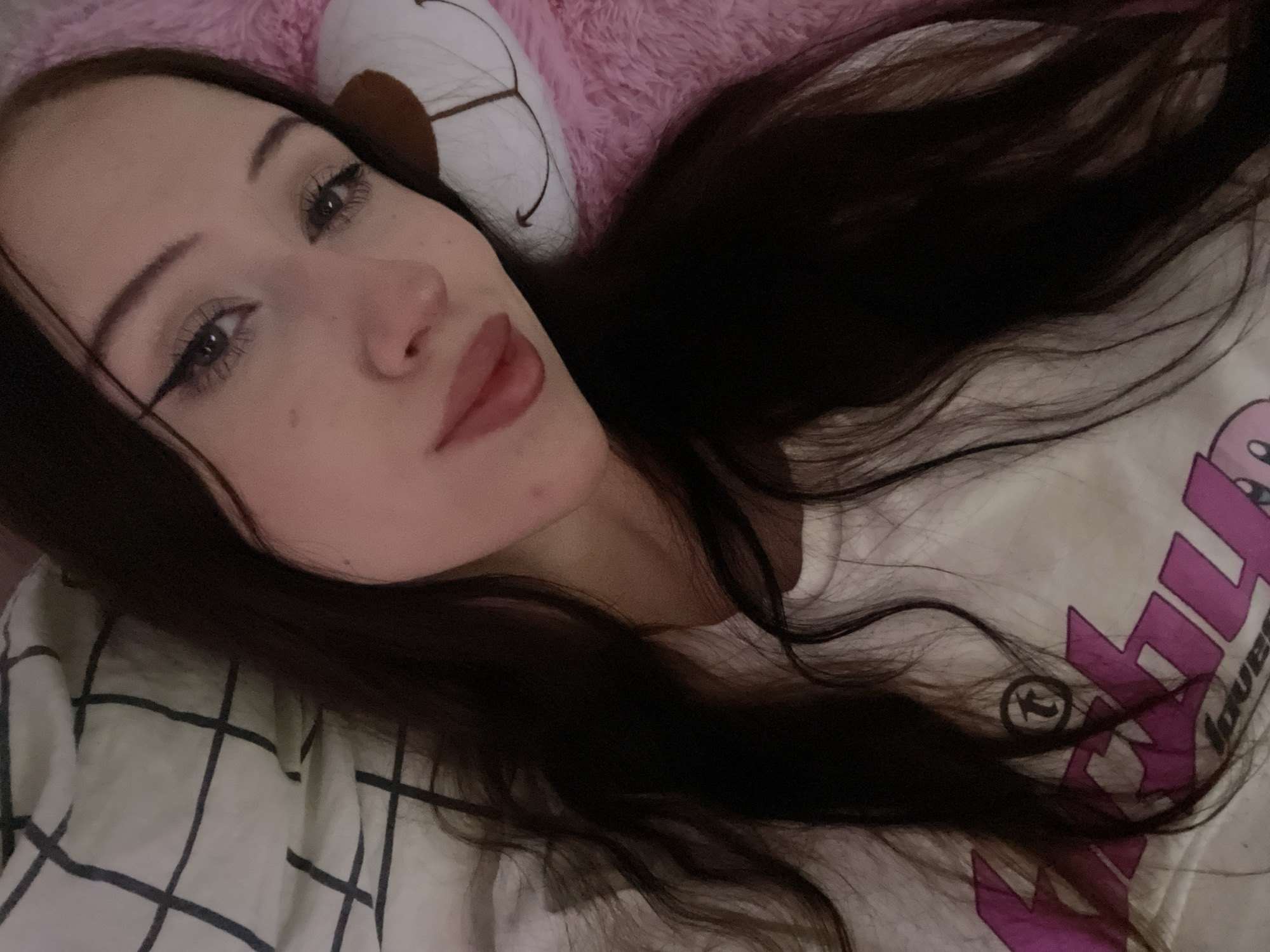 The image shows a close-up selfie of a young woman with long, dark hair. She is lying down, with her head resting on a pink, fluffy object that appears to be a stuffed animal. A white and dark-lined checkered blanket is visible behind her. The woman is wearing a white t-shirt with a pink graphic design on it. She has makeup on, including winged eyeliner and lipstick. The lighting suggests the photo was taken indoors, possibly in a bedroom. There is no information in the image to suggest it is located in Votkinsk, Russia.