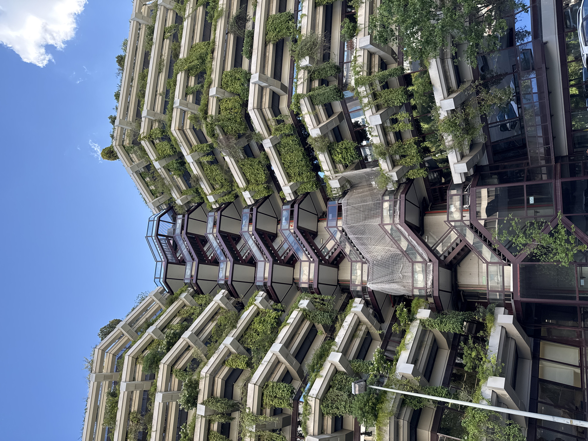 The image captures a unique architectural structure located in Barcelona, Spain. It showcases a multi-story building with a distinctive design featuring tiered, angular concrete balconies adorned with lush greenery. The building's façade is characterized by a zig-zag pattern formed by the projecting balconies, creating a visually striking effect. The concrete structure is interspersed with glass panels, and brown metal framing accents the design. The abundant vegetation cascading from the balconies adds a natural element to the modern construction. A clear blue sky is visible in the background, contrasting with the building's angular form.