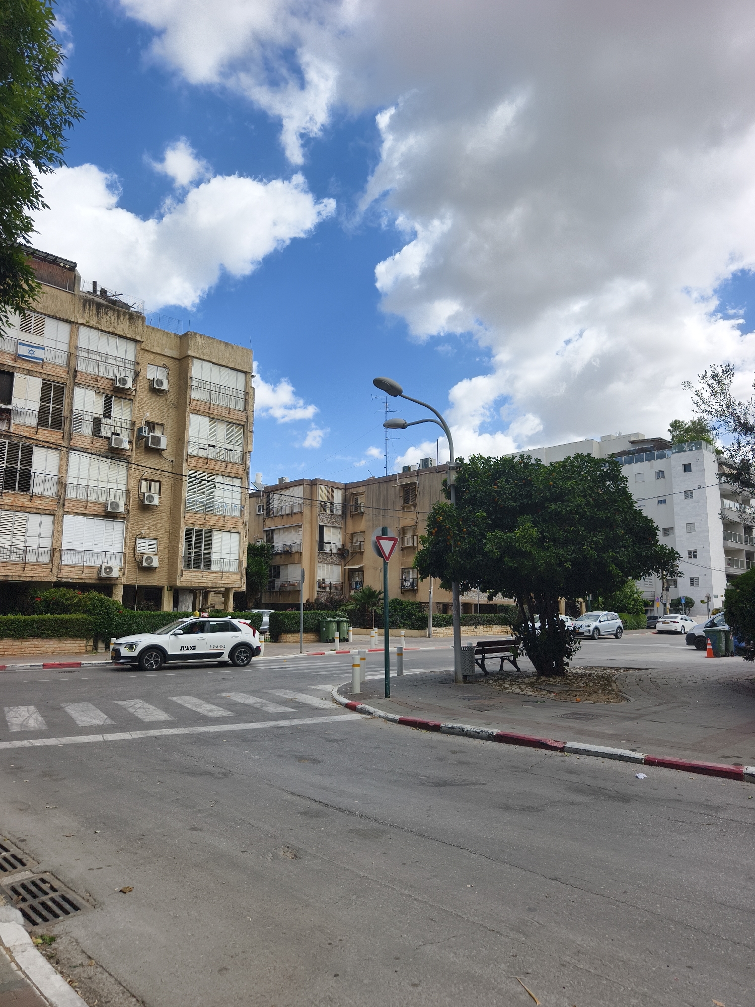 The image depicts a street scene in Petah Tikva, Israel. The buildings are old, multi-story structures with balconies, air conditioning units, and some are adorned with the Israeli flag. A white car with Hebrew text and the number 19634 is crossing a crosswalk. There is a yield sign on a pole near a tree with green foliage and orange fruit, next to a street light. A bench sits near the tree. There are other cars parked along the street and a white apartment building is in the background. The sky is partly cloudy.
