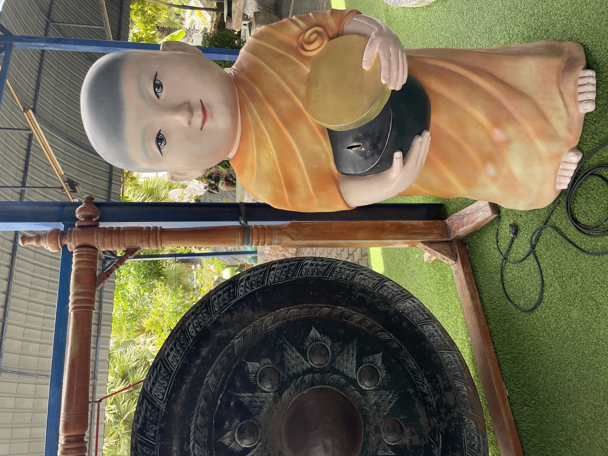 The image captures a life-sized statue of a young monk standing on a green, artificial turf floor, likely in Bang Sare, Thailand. The monk is depicted with a shaved head, large eyes, and a gentle smile, wearing an orange robe. In his hands, he holds a dark alms bowl topped with a golden lid. To the left of the statue is a large, dark gong suspended from a wooden frame. The background reveals a covered outdoor area with palm trees and other greenery visible beyond a blue metal support beam. A black electrical cord lies on the ground near the base of the statue.