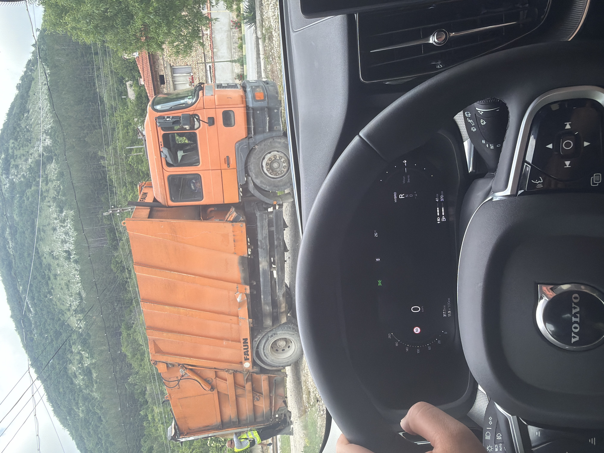 The media file presents a first-person perspective from inside a Volvo car, looking out at an orange garbage truck in a rural or semi-urban setting, specifically Troitsa, Bulgaria. A hand is visible holding the car's steering wheel on the right.
The car's interior features a black "VOLVO" branded steering wheel and a digital dashboard. The dashboard indicates an outside temperature of 26°C, the car is stationary (0 km/h), with a remaining range of 370 KM. A trip odometer shows 317.1 KM, and average fuel consumption is 11.9 L/100KM. The gear selection area displays "HYBRID" and "RND". Various controls for lights ("AUTO") and wipers ("INT") are also visible.
Outside the car, a large, bright orange garbage truck is the central subject. The brand name "FAUN" is clearly visible on its side. A person wearing a high-visibility vest is seen near the rear of the truck, suggesting ongoing work or a stop. The background is dominated by a large, green, tree-covered mountain or hillside under an overcast sky. Power lines are strung across the landscape. Part of a house with a red-tiled roof and brickwork is visible to the right, indicating a residential area. The scene depicts a warm, possibly summer, daytime encounter with a waste collection vehicle.