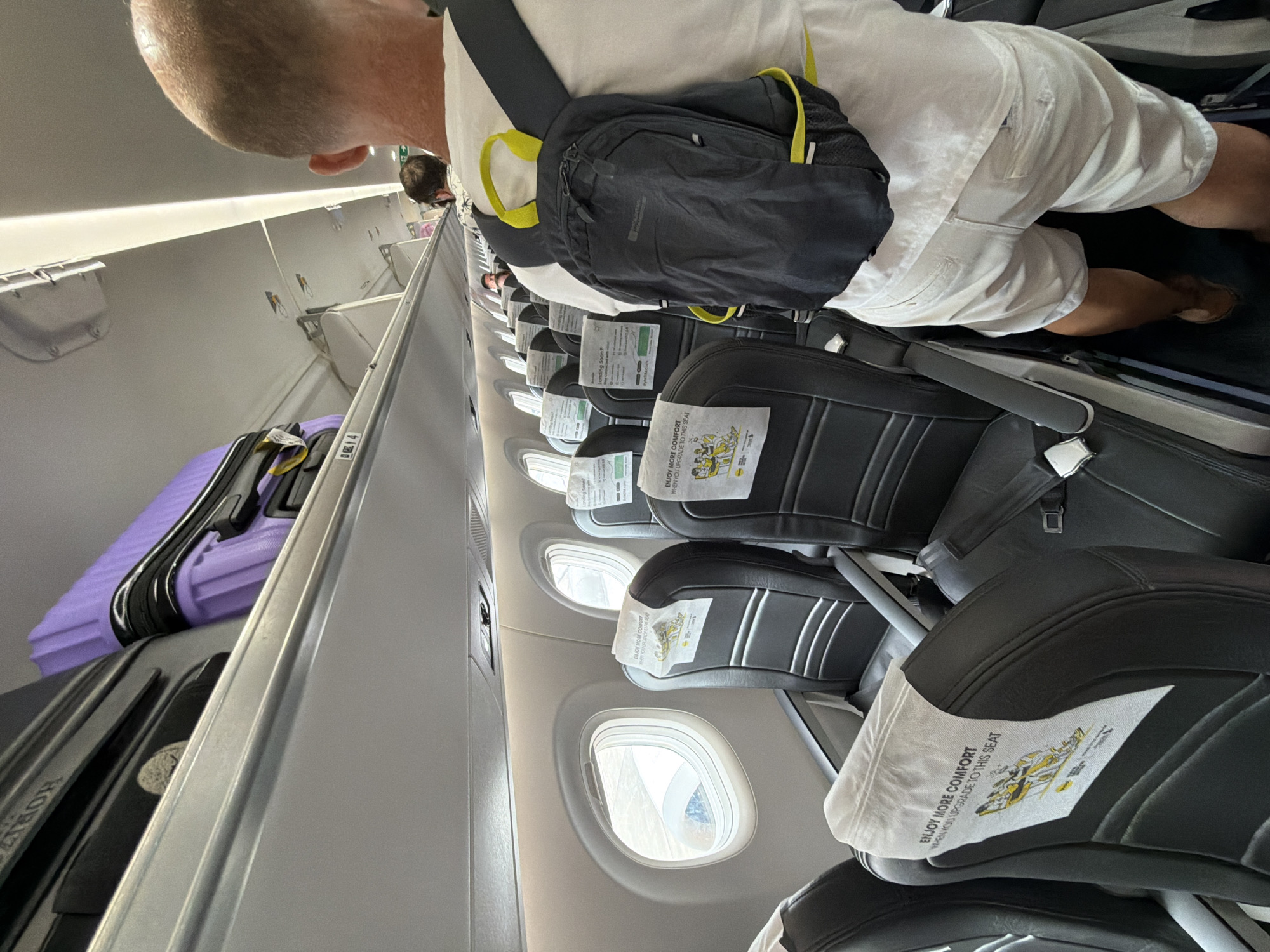 The image depicts the interior of an airplane cabin, likely on an airline serving Thailand, given the mention of Ko Samui District. In the foreground, a man with a bald head and fair skin is viewed from behind, wearing a white t-shirt and light-colored shorts, with a dark backpack with yellow accents on his back. He is standing in the aisle of the plane, facing forward, and appears to be looking towards the front of the cabin.
The cabin features rows of black airplane seats with white headrest covers that have printed graphics and text. Some of these headrest covers display the phrase "ENJOY MORE COMFORT WHEN YOU UPGRADE TO THIS SEAT." The seats are arranged in a typical 2-2 configuration. Overhead compartments are visible, with luggage, including a purple suitcase and a dark grey suitcase, stored inside. The windows of the airplane are visible, showing a bright, possibly daytime, exterior. The lighting in the cabin is a mix of overhead lights and natural light from the windows. The overall impression is a typical scene of passengers boarding or seated on a commercial flight.