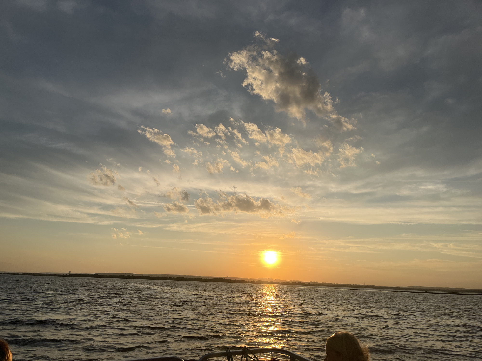 The image depicts a serene sunset over a body of water, likely a bay or large lake, in the city of Shumeika, Russia. The sun, a bright orb, is positioned low on the horizon, casting a warm, golden glow across the scene. Its reflection shimmers on the rippling surface of the water, which occupies the lower half of the frame. The sky above is a canvas of soft hues, transitioning from the vibrant orange of the sunset to a dusky blue, with scattered clouds tinged by the fading light. A silhouetted landmass, possibly the Shumeika coastline, forms a distant horizon. In the foreground, the edge of what appears to be a boat or railing is visible, with a person's blonde hair and shoulder peeking into the frame from the right. The overall atmosphere is one of tranquility and natural beauty.