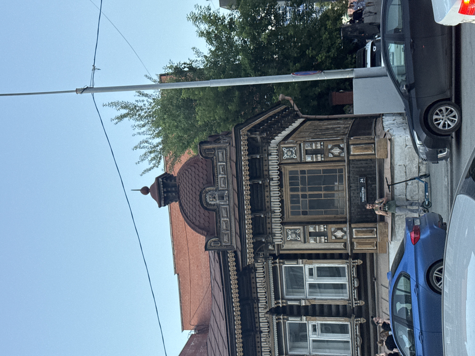 The image captures a view of a historic wooden house on Tolmacheva Street in Yekaterinburg, Russia, characterized by intricate carvings and a domed roof. In front of the house, a man is standing on a blue electric scooter, holding a phone, and looking at it. To the left, a blue car is parked, and to the right, a dark-colored car is parked. Several people are visible walking in the background. The sky is clear and blue, suggesting it is daytime. The architecture of the house is a prominent feature, with detailed wooden decorations on its facade. The surrounding environment includes trees and a streetlamp.