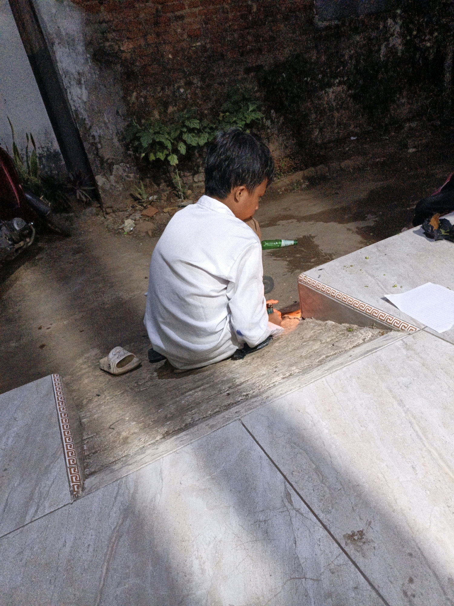 An undated, eye-level, outdoor, vertical medium shot captures a young boy, viewed from behind, sitting on a textured, grey concrete surface, possibly in Tanahbaya, Indonesia. The boy, wearing a white long-sleeved shirt, is hunched over, seemingly engaged with something on the ground. A single flip-flop lies to his left. To his right, a green bottle and a silver object, possibly a lighter, are visible on the ground. The background consists of a weathered, dark brick wall with patches of green foliage, and to the far left, a dark motorcycle with red accents and some foliage can be seen. A light-colored, raised concrete structure with decorative trim runs diagonally across the foreground and right side of the image, with a sheet of white paper placed on it. Sunlight casts shadows across the scene, creating areas of light and dark.