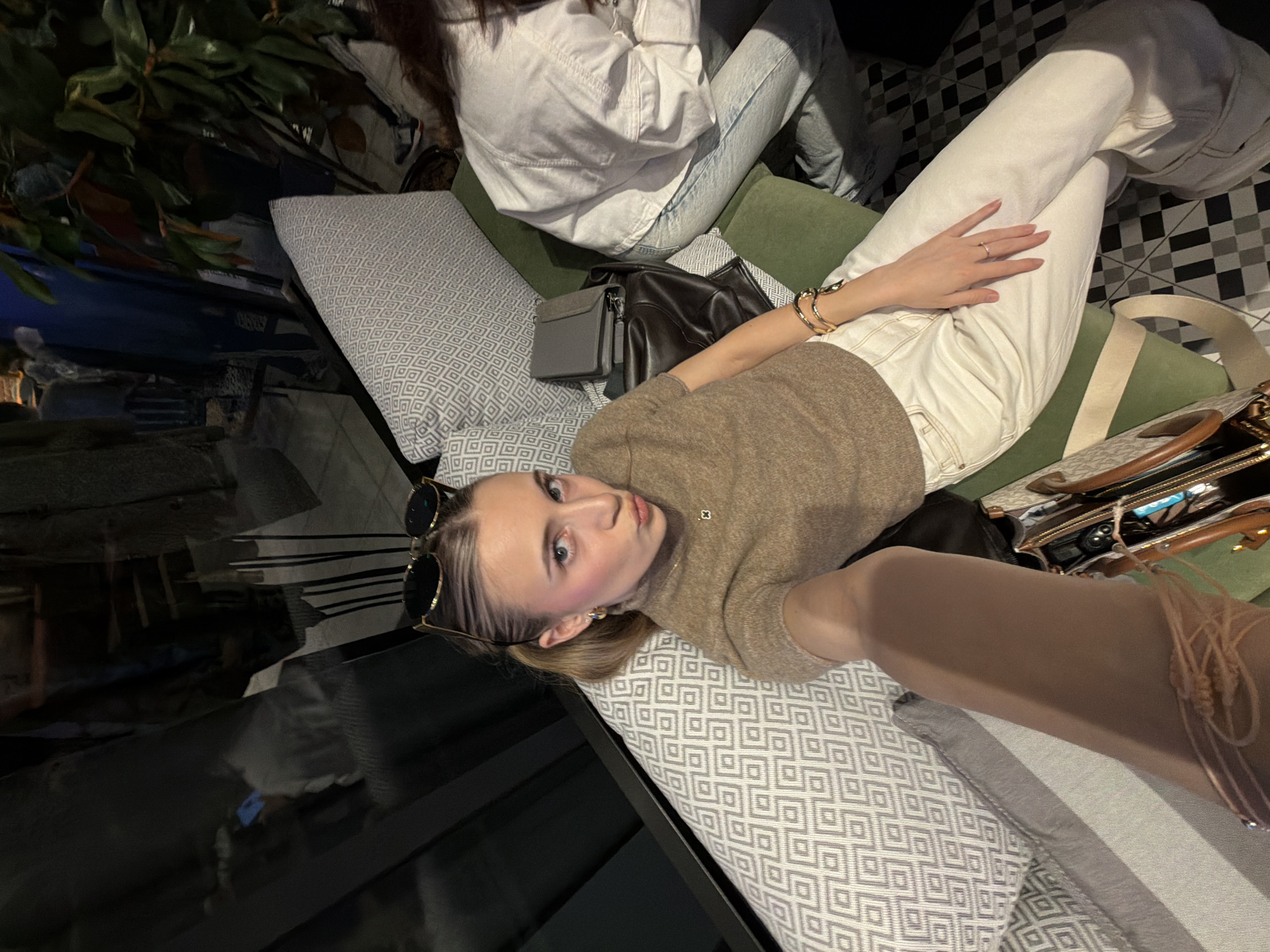 The image presents an indoor scene featuring two individuals within a commercial establishment in Plovdiv, Bulgaria. A young adult female is positioned centrally, capturing a selfie. She has light-colored hair styled into two low ponytails, sunglasses resting on her head, and is wearing a short-sleeved, light brown knitted top, light-colored high-waisted pants, gold hoop earrings, a small clover pendant necklace, and multiple gold bracelets on her right wrist. Her left hand, adorned with a thin ring on the ring finger, is visible resting on her leg. Her lips are pursed. She occupies a green-cushioned bench or sofa accented with light grey patterned throw pillows. A dark brown handbag and a grey wallet are placed on the seating surface beside her. To her right, another individual is partially visible, wearing a white jacket and light blue jeans, seated on a similar green-cushioned surface. The background features a dark reflective surface to the left, displaying ambient reflections. To the right, a large green potted plant is present. The floor is covered in a black and white checkerboard tile pattern. The overall lighting is dim, consistent with an evening or atmospheric indoor setting. The specific location within the city of Plovdiv is not identifiable from the visual evidence.