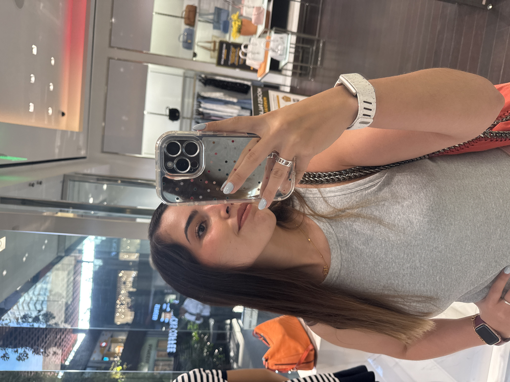 A woman is taking a selfie in what appears to be a shopping mall. She is holding a cell phone in her right hand, wearing rings and a white smart watch. She is wearing a gray ribbed tank top and has a chain necklace. A red-orange handbag is visible near her right shoulder. The background features storefronts and displays, including purses and clothing. Reflections of the outside environment, including trees and buildings, are visible in the windows. The city can be identified as Puebla, Mexico because the exterior reflection contains 'CIR COFFEE' which refers to the coffee shop chain Círculo de Café, a Mexican coffee shop chain with several locations in Puebla, Mexico.