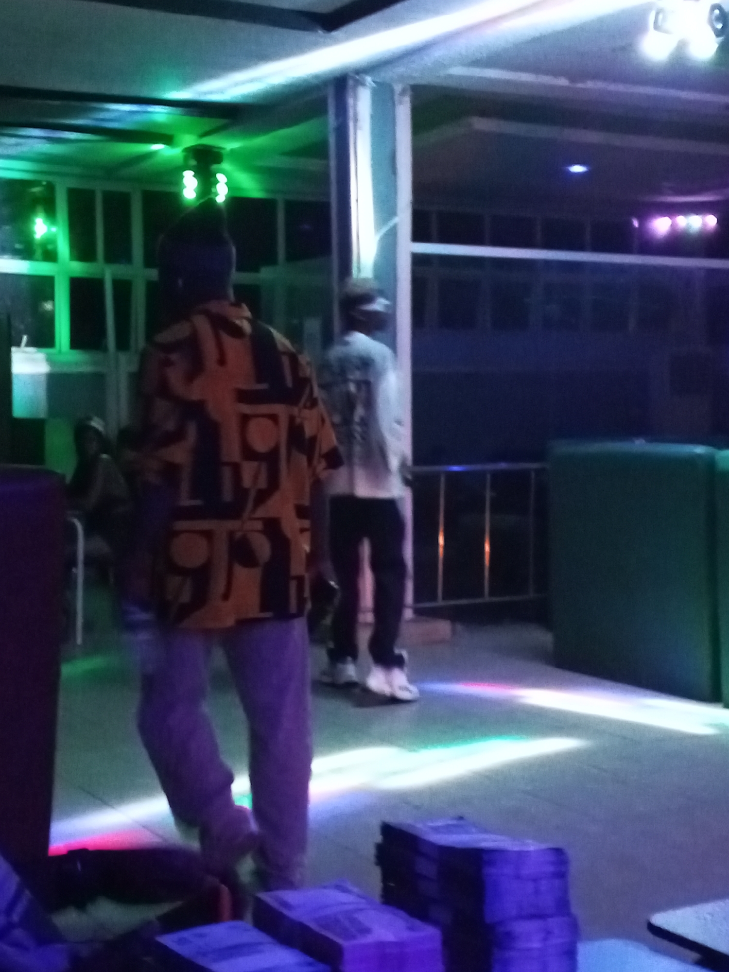 The image depicts an indoor setting, likely a club or lounge, characterized by dim lighting and the presence of colorful lighting effects. Two male figures are visible, both facing away from the camera. The individual in the foreground is wearing a patterned orange and black shirt paired with light-colored pants, and is in motion. The second individual, further in the background, is wearing a white shirt and dark pants, and is standing still.
The environment features a combination of seating areas and open space. In the background, there is a balcony-like structure with metal railings and square-paneled windows. Green cushioned seating is positioned to the right of the frame. In the foreground, there are stacked objects of a dark color. The floor is reflecting the multicolored lights, contributing to the overall ambiance. The location is identified as Unknown, Unknown.