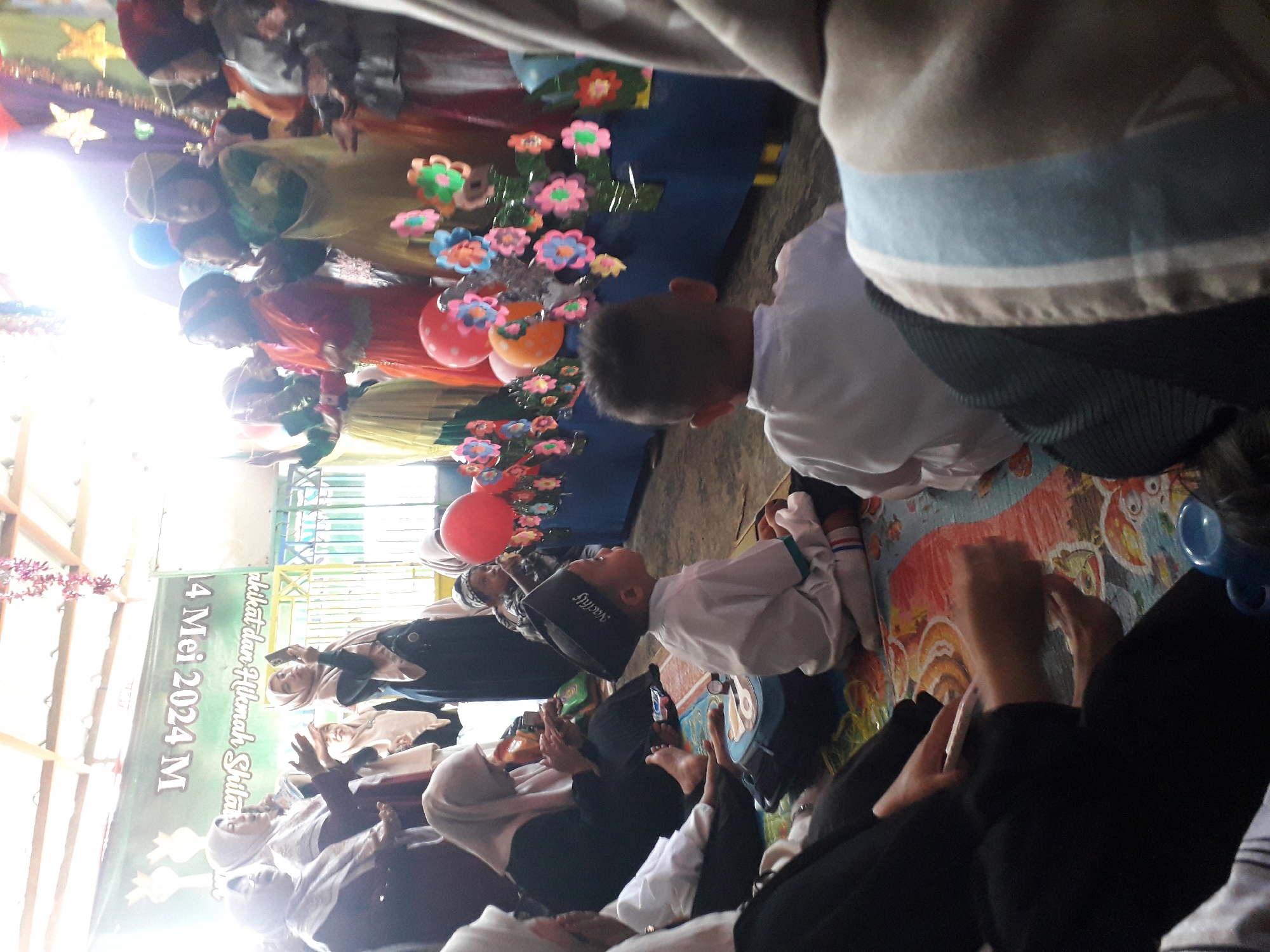 This is a medium shot taken from a low angle of a gathering, likely a religious or cultural event, with many people, predominantly women and children, present. The scene appears to be indoors, with a wooden ceiling visible and what looks like a large banner hanging in the background. The banner has text in a foreign language and a date that appears to read "4 Mei 2024 M". The participants are dressed in traditional attire, with many women wearing hijabs and long dresses. Some children are seated on the floor, facing a stage-like area decorated with colorful paper flowers and balloons. One child in the foreground wears a black cap and white clothing, facing to the right of the frame. Another child, in the mid-ground and facing away, also wears white. Several women in the background are looking towards the stage, with some appearing to be taking photos with their phones. The overall atmosphere seems festive and communal. The context provided mentions the city of Cankuang, Indonesia.