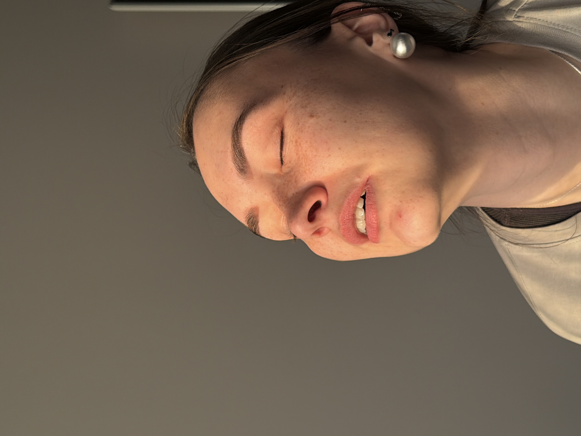A close-up, rotated image depicts a fair-skinned person with freckles and closed eyes. The individual has dark hair pulled back, visible behind the ear which is adorned with a large, spherical, silver earring. The person's mouth is slightly open, revealing teeth. The individual wears a light-colored garment. The background is a plain grey. There is no verifiable evidence to place this image in Chaikovsky, Russia.