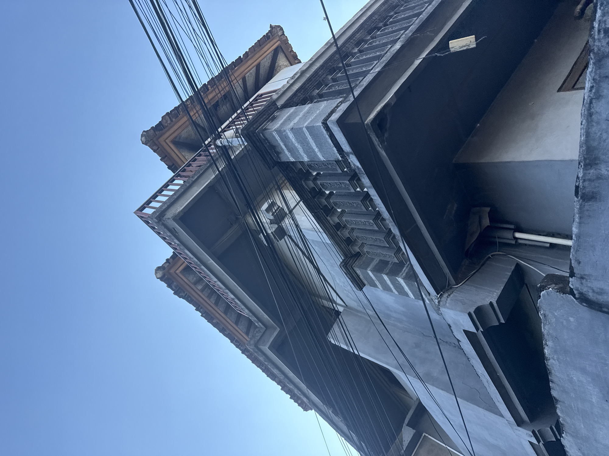 The image shows an upward-angle shot of the corner of a multi-story building against a clear blue sky. The building has a gray concrete facade with decorative elements, including a balustrade with carved panels on the upper level. The roofline is visible, with terracotta tiles and wooden trim. A dense network of black electrical wires runs across the upper portion of the image, crossing over the building and appearing to be suspended by poles or other structures not fully visible. The lower part of the image shows a gray concrete wall or ledge. The overall impression is of an urban setting in Denpasar, Indonesia, characterized by its architecture and ubiquitous electrical infrastructure. There are no people or overt actions depicted.