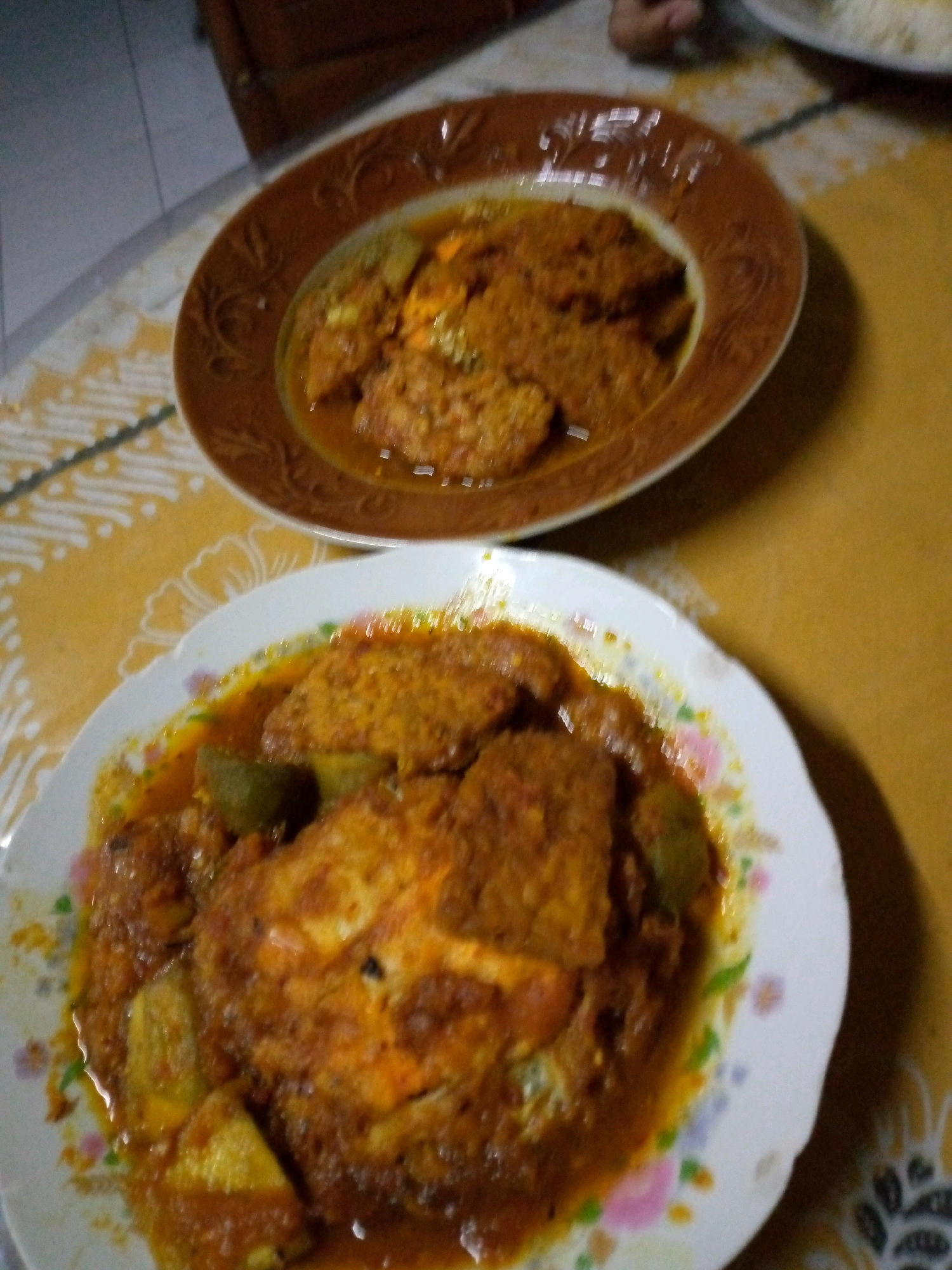The image shows two plates of Indonesian food on a yellow tablecloth. The plate in the foreground is white with a floral pattern and contains a dish with a reddish-brown sauce, pieces of what appears to be fish or meat, and some green vegetables. The plate in the background is brown with a decorative rim and contains a similar dish with a reddish-brown sauce. The setting appears to be a home or casual dining environment, possibly in Sumberwono, Indonesia. There are no people visible in the image. The lighting is dim, suggesting it might be evening or indoors.