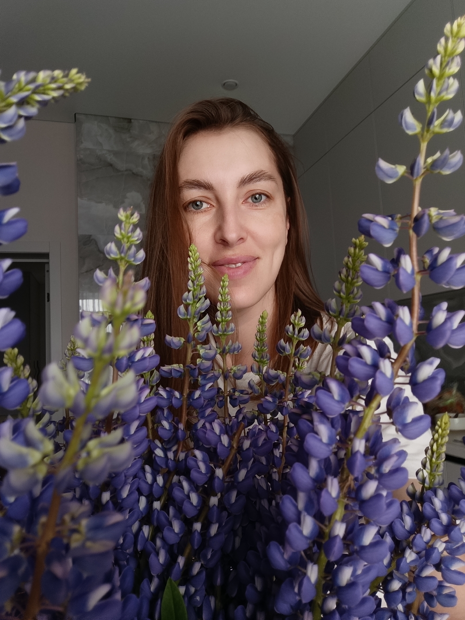 A woman with shoulder-length brown hair and fair skin smiles directly at the camera from behind a large bouquet of purple lupine flowers. The lupines frame her face, partially obscuring her body. She wears a white top. The background suggests an interior setting, possibly a modern apartment, with light gray walls and what appears to be a door on the left side. The overall lighting is soft and natural. The location is not discernible from the image.
