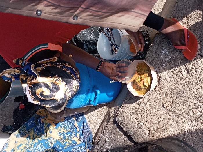 This image depicts an outdoor food service scene in Jos, Nigeria, under bright sunlight. A person, whose head is covered by a black headwrap with gold and white patterns and wearing a red top, is observed serving food. Their dark-skinned hands are centered, with the left hand holding a clear plastic lid over a white, polygonal disposable container filled with a greenish-yellow stew containing solid components. The right hand holds a small, clear plastic container filled with reddish-orange powdered seasoning, positioned to be added to the meal. The individual's right foot, clad in an orange flip-flop, rests on the cracked, grey concrete ground, which exhibits harsh shadows. To the left, a large blue plastic bucket is visible. To the right, a stack of white bowls holds numerous metal spoons and forks. In the background, blue and yellow patterned fabric is partially visible, alongside other dark materials.
