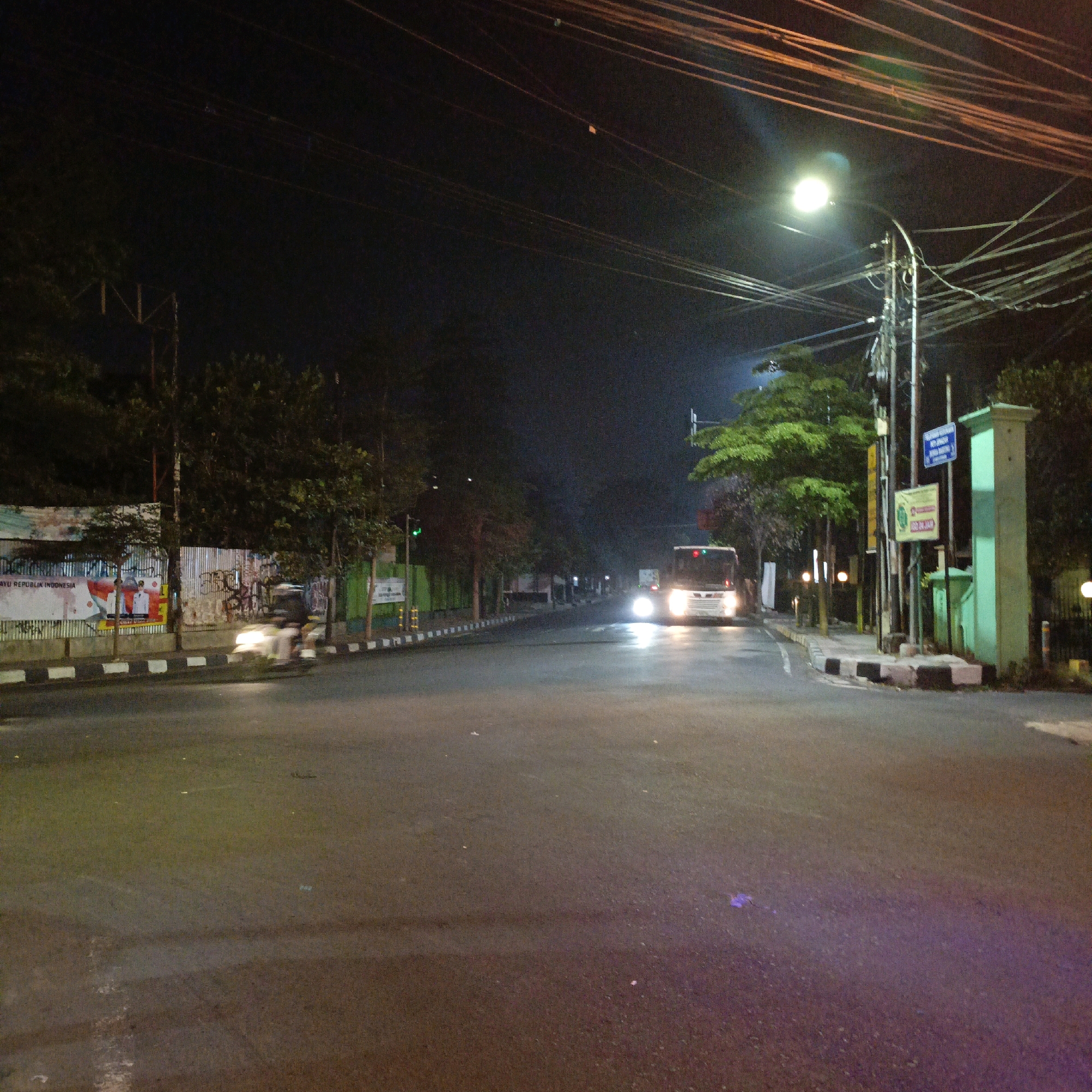 The image depicts a nighttime street scene in Salatiga, Indonesia. A white bus with its headlights on is traveling down the center of the asphalt road, which is wet and reflects the light. The road is lined with sidewalks and trees, and a traffic light is visible on the left side of the image, showing a green signal. Power lines are strung overhead, supported by poles, and a street lamp illuminates the scene from the right. A motorcycle with a rider, blurred due to motion, is moving on the left side of the road. There are advertisements and graffiti on a corrugated metal fence behind the sidewalk on the left. The overall atmosphere is one of a typical urban street at night, with visible traffic and street infrastructure.