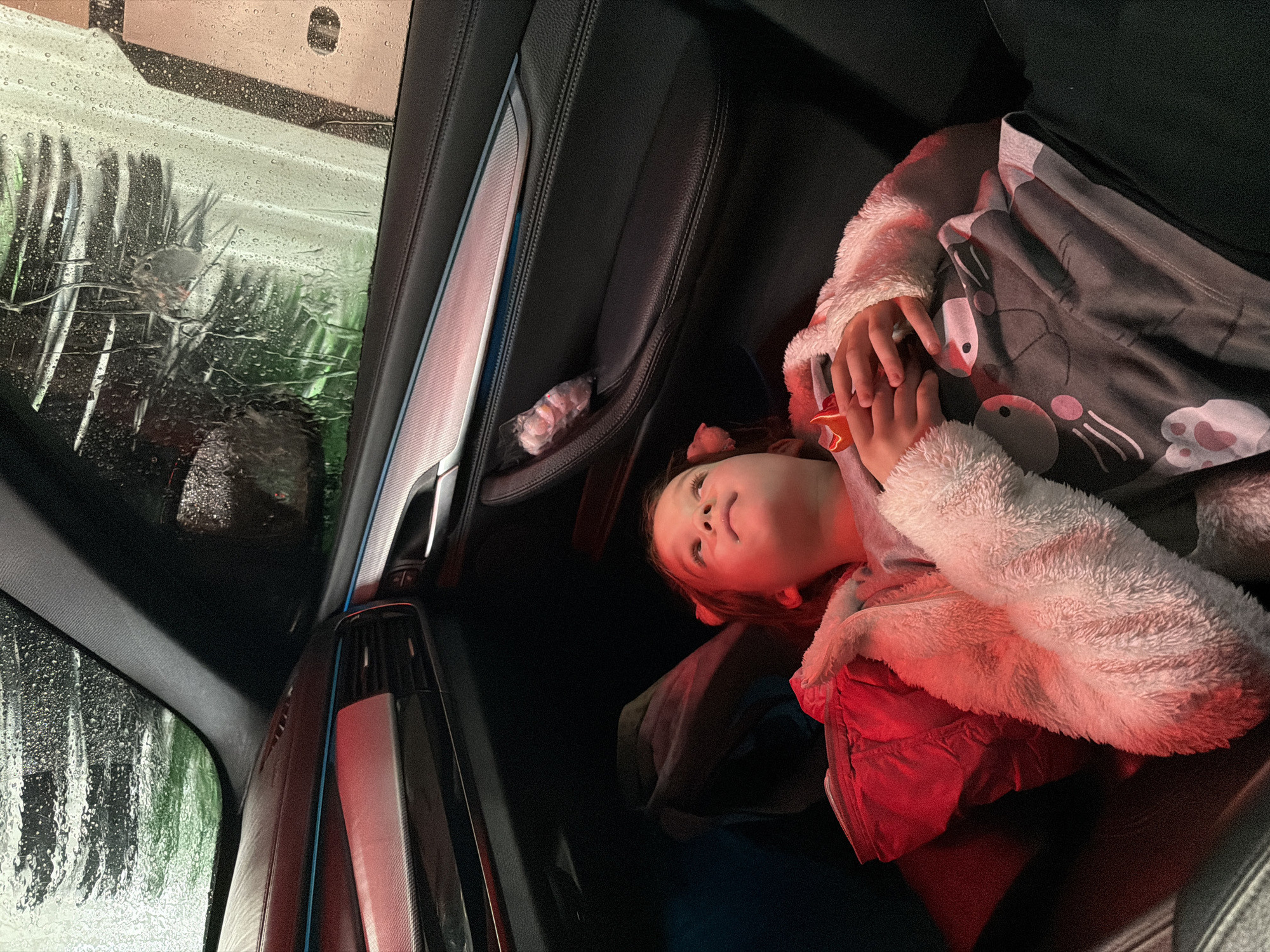 A young child, wrapped in a fuzzy white jacket and covered with a grey blanket printed with a cartoon animal design, is seen inside a car during a car wash. The car wash brushes and soapy water are visible through the car window. A small wrapped item is in the door storage compartment. The interior of the car appears to be dark, modern, and possibly leather. The location cannot be determined from this image.