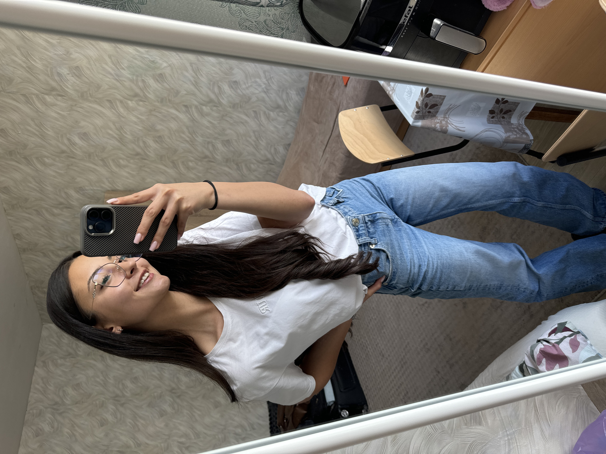 The image, originally rotated clockwise, presents a mirror selfie of a young woman standing in an indoor setting, likely a home in Sofia, Bulgaria. She has long, dark hair cascading over her shoulder and is wearing round-framed glasses, a white short-sleeved t-shirt, and blue denim jeans. Her left hand rests on her hip, while her right hand holds a smartphone with a black carbon-fiber-like case, positioned to capture her reflection. She has a gentle smile, looking towards the phone's screen. A black elastic hair tie is visible on her left wrist, and her fingernails are painted a light pink.
The reflected background reveals a light-colored wall with a wavy, textured wallpaper. Towards the right, a light wooden chair with a curved seat and back is visible, along with a table adorned with a patterned runner featuring brown and grey leaf designs. An appliance, identified by the visible text "Tefal Easy Fry & grill," sits on a surface behind the table. The floor appears to be covered with a textured rug or carpet. On the left chest of the woman's white t-shirt, a white, stylized logo in a gothic-style font is discernible. The room is brightly lit, suggesting it is daytime.